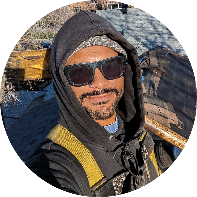Roofing MTL employee outdoors wearing sunglasses, a gray beanie, a black hoodie, and a yellow safety strap, smiling and holding a long wooden tool, with a background of rooftops and trees.
