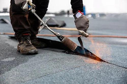 A worker using a torch to apply hot asphalt onto a flat roof in Montreal.