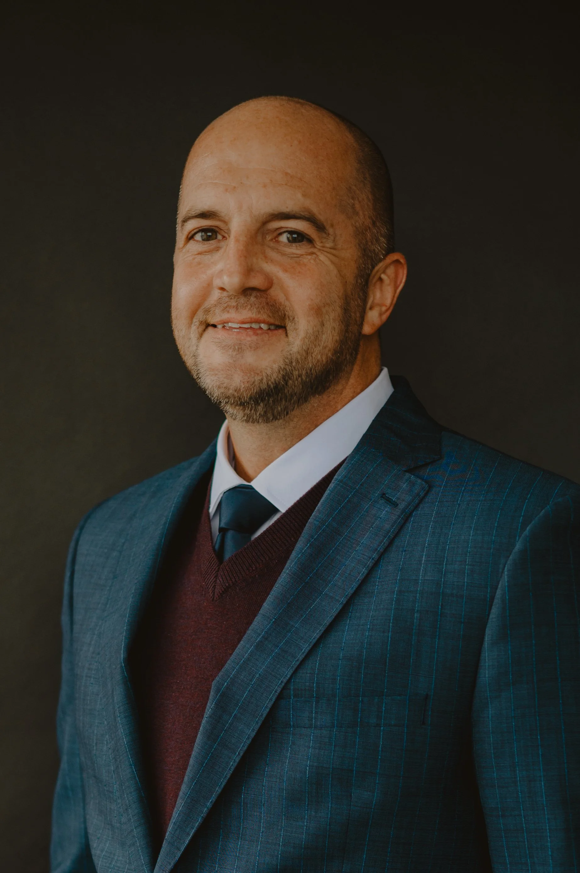 Headshot of a man in a suit and tie, smiling against a dark background.
