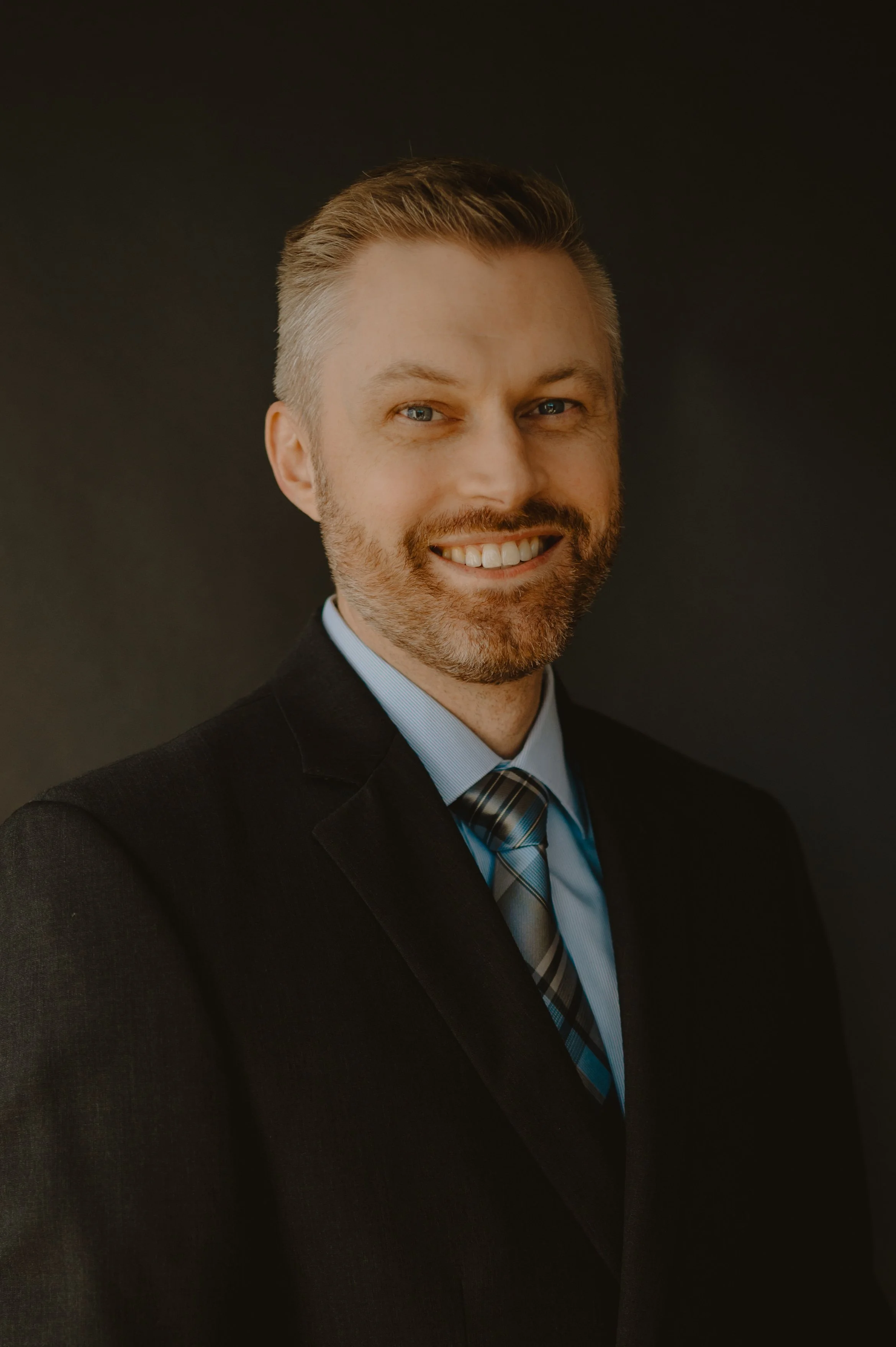 Headshot of a smiling man with light skin, short light brown hair, a beard, wearing a dark suit, light blue shirt, and tie, against a dark background.