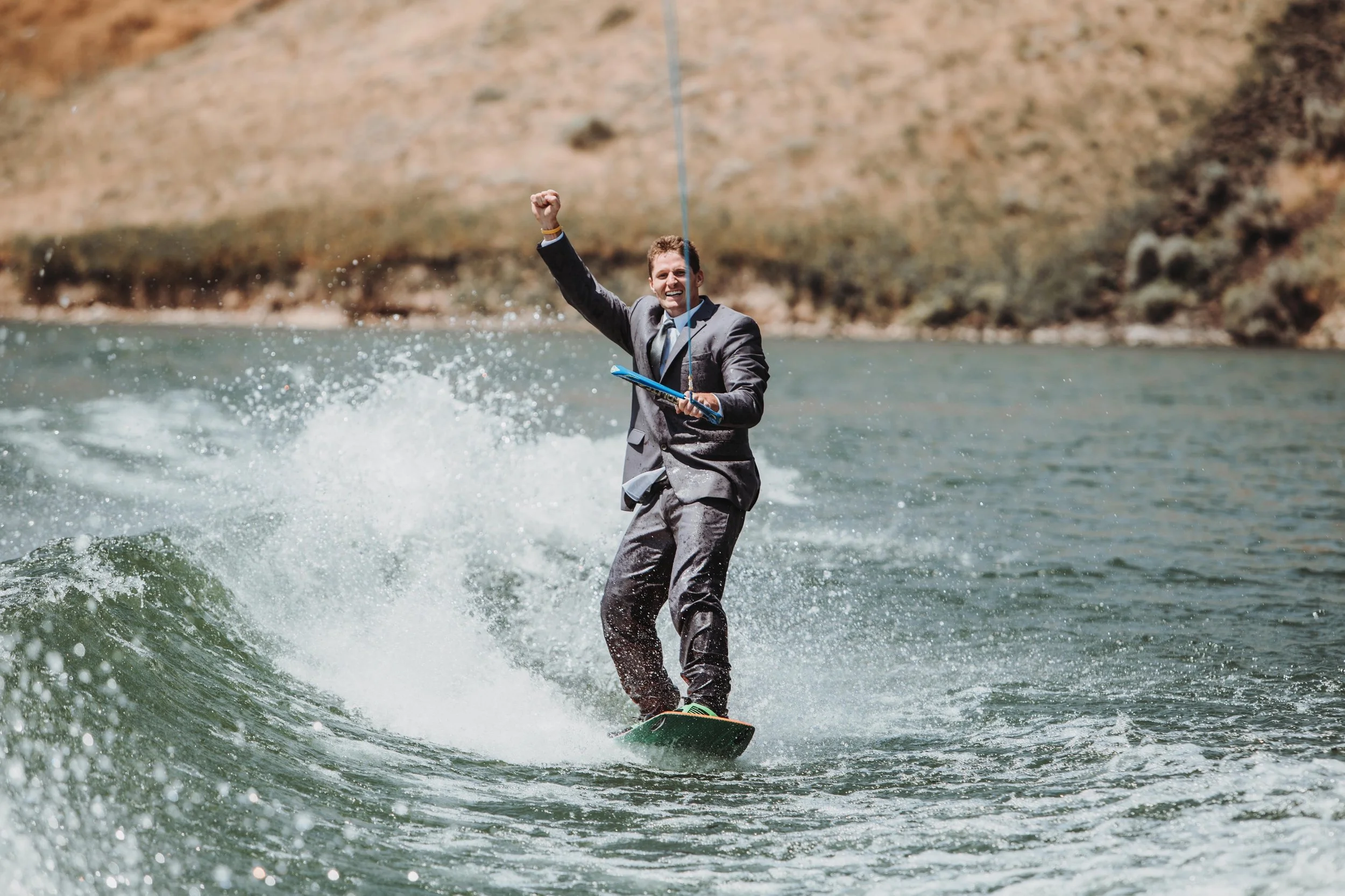 A man dressed in a business suit riding a wakeboard on a body of water, holding a fishing rod with a line in the water, smiling and raising one arm in triumph.