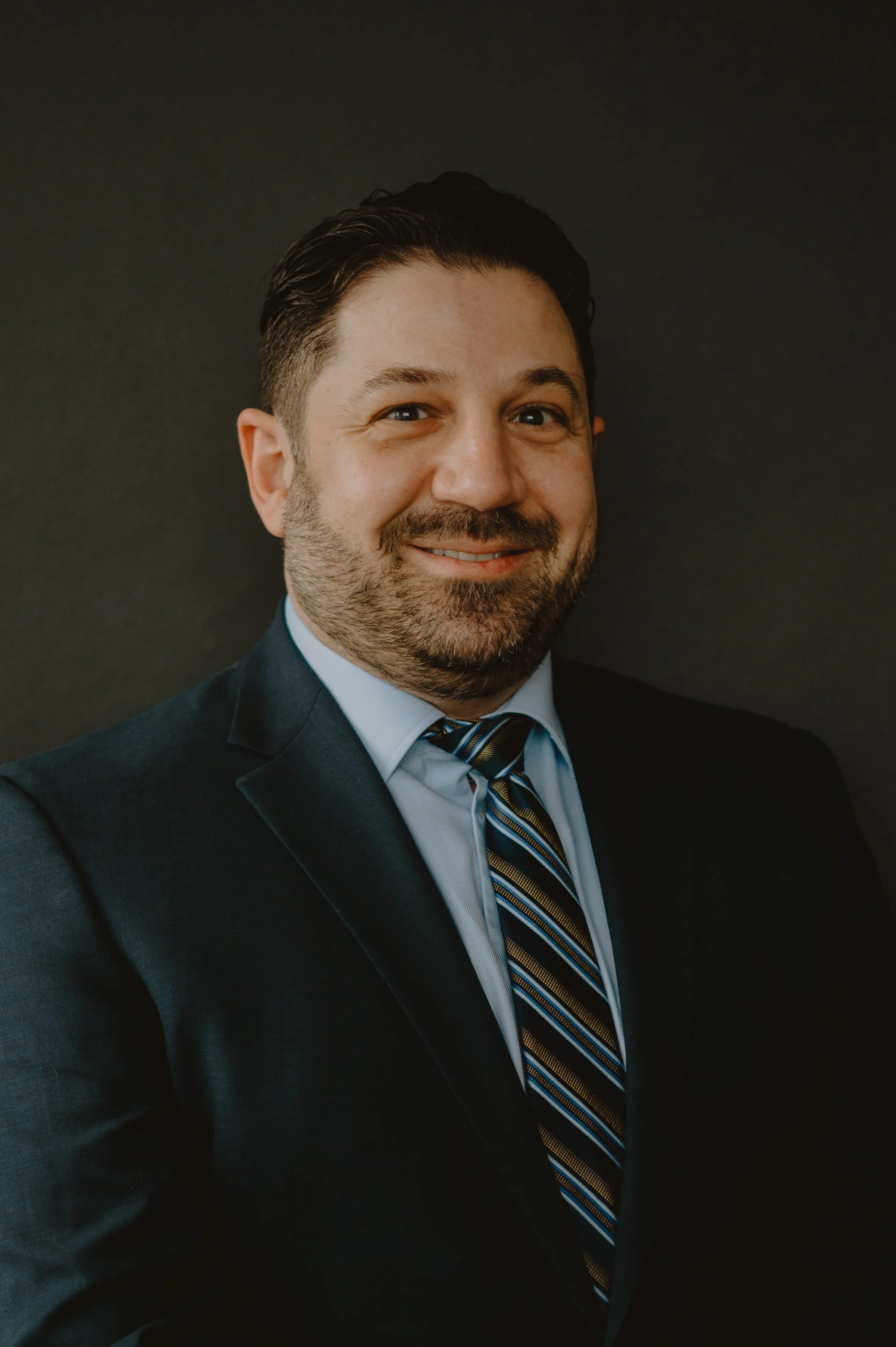 A headshot of a man smiling, wearing a dark suit, light blue shirt, and a striped tie, against a dark background.
