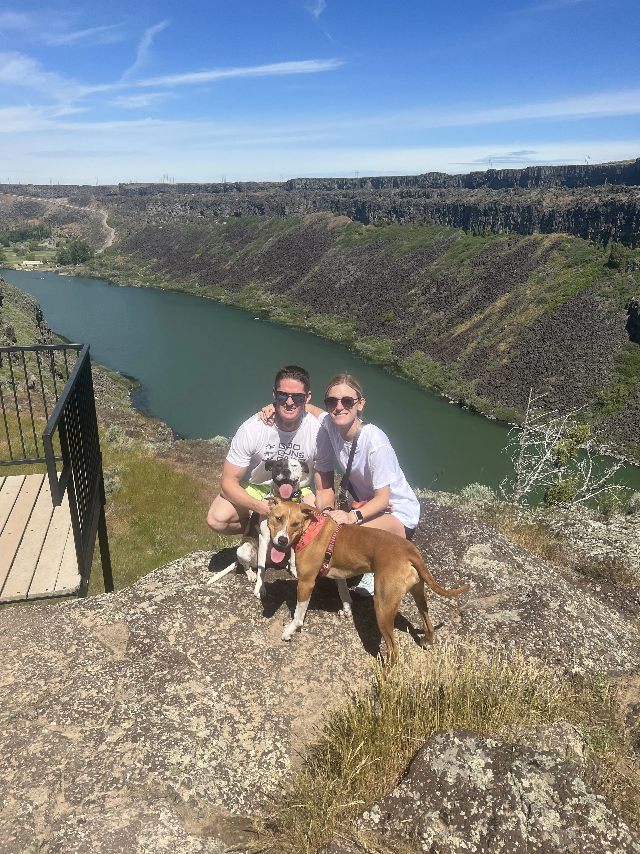 A couple smiling with their two dogs on a rocky overlook with a river and steep cliffs in the background on a sunny day.