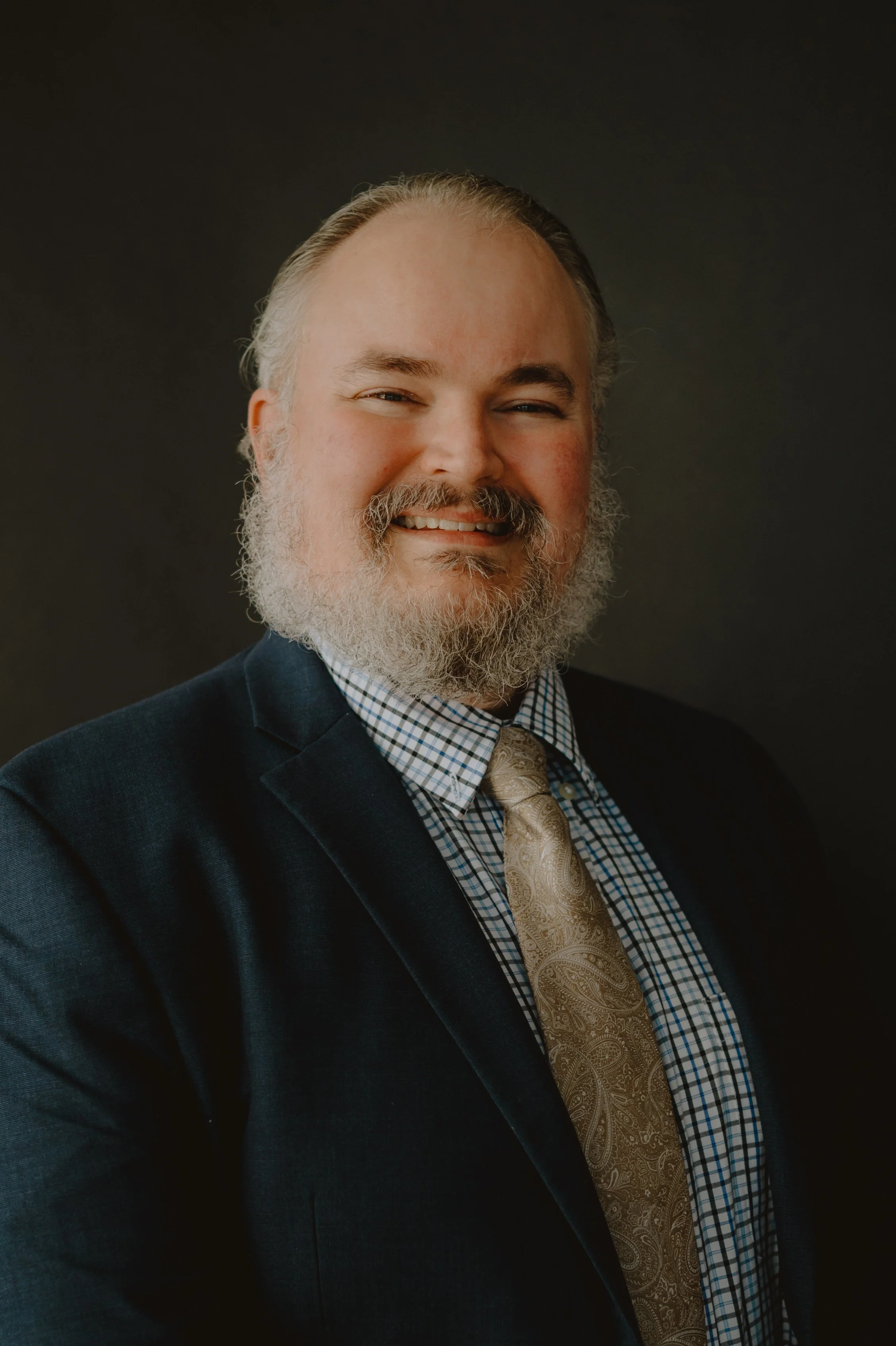 A man with gray hair, beard, and mustache, smiling, dressed in a checkered shirt, paisley tie, and dark blazer, against a dark gray background.