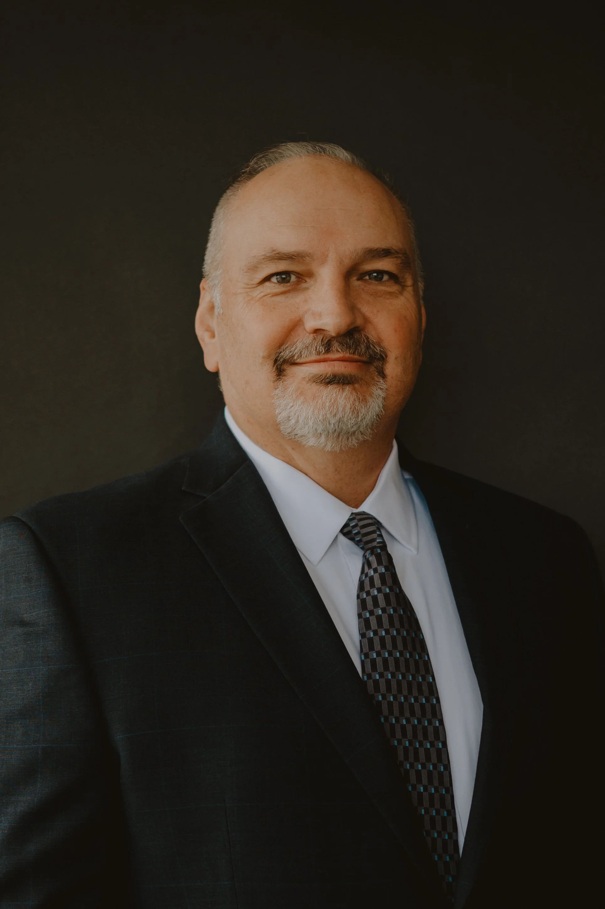 Portrait of a middle-aged man with gray hair and beard wearing a dark suit, white shirt, and patterned tie standing against a dark background.
