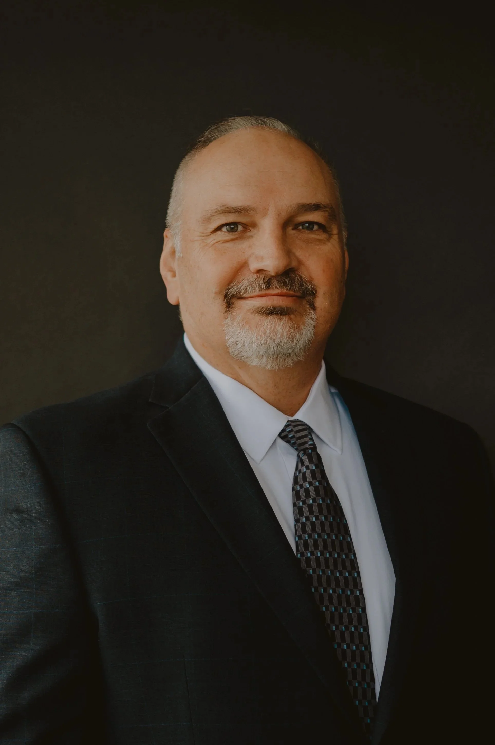 Portrait of a middle-aged man with gray hair and beard, dressed in a dark suit, white shirt, and patterned tie, standing against a dark background.