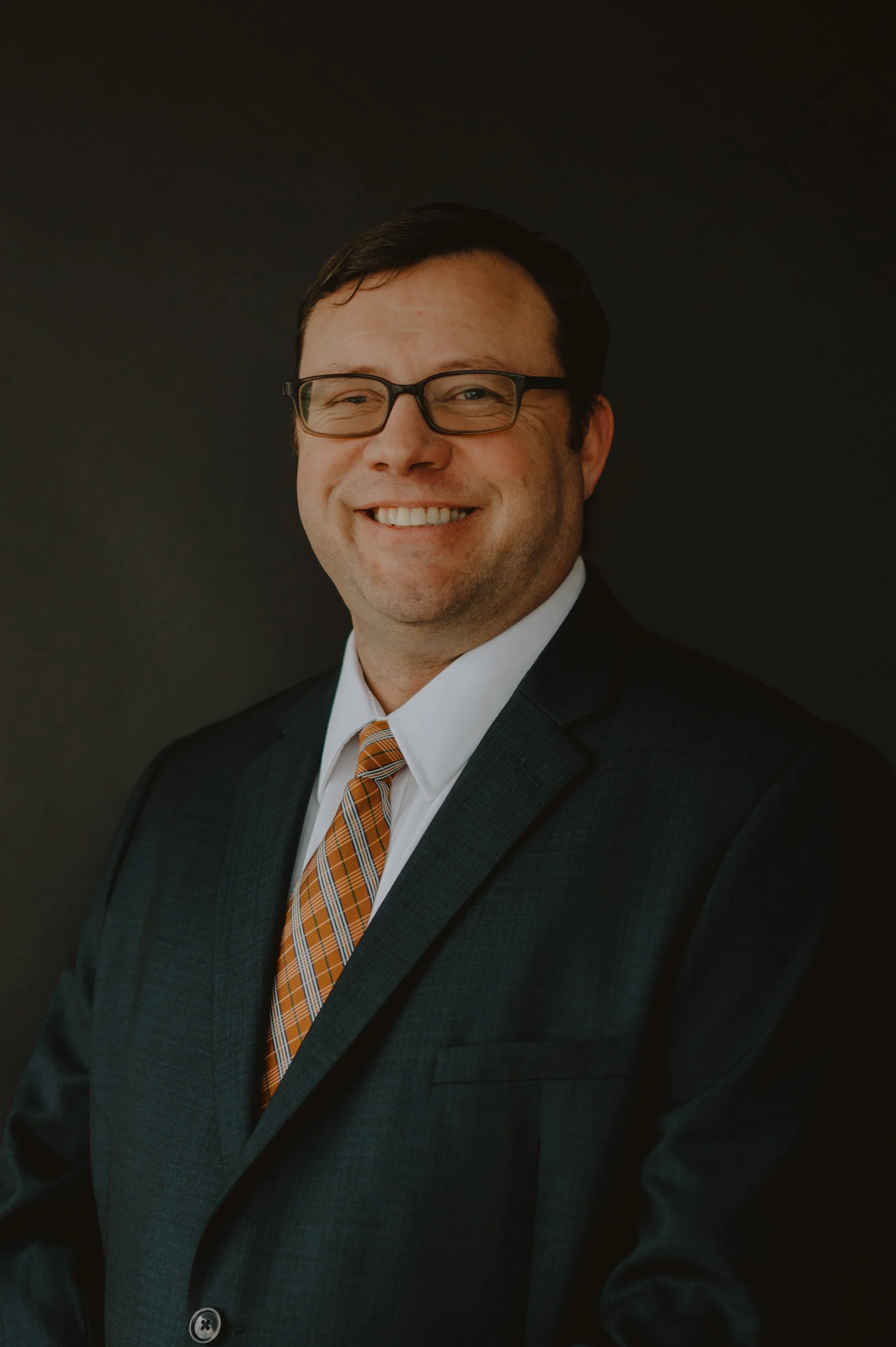 Professional headshot of a man wearing glasses, a dark suit, a white shirt, and an orange plaid tie, smiling against a dark background.