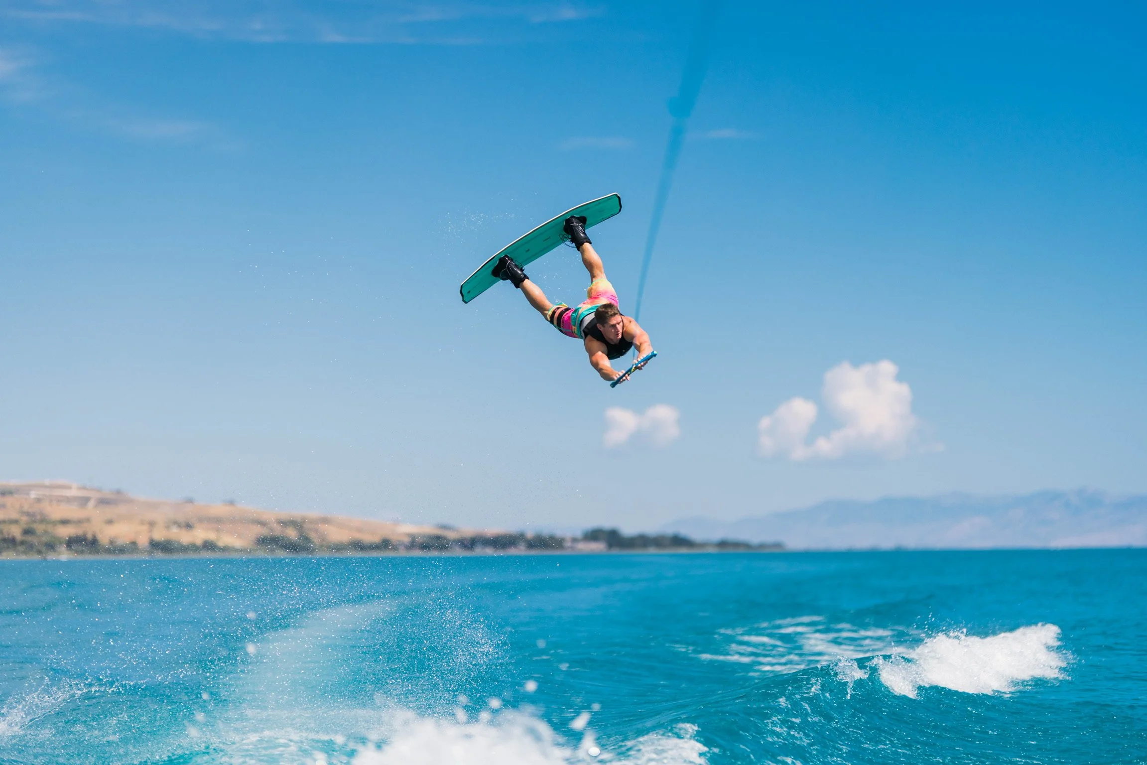 Person wakeboarding in the water, mid-air, with a clear blue sky and distant shoreline and mountains in the background.