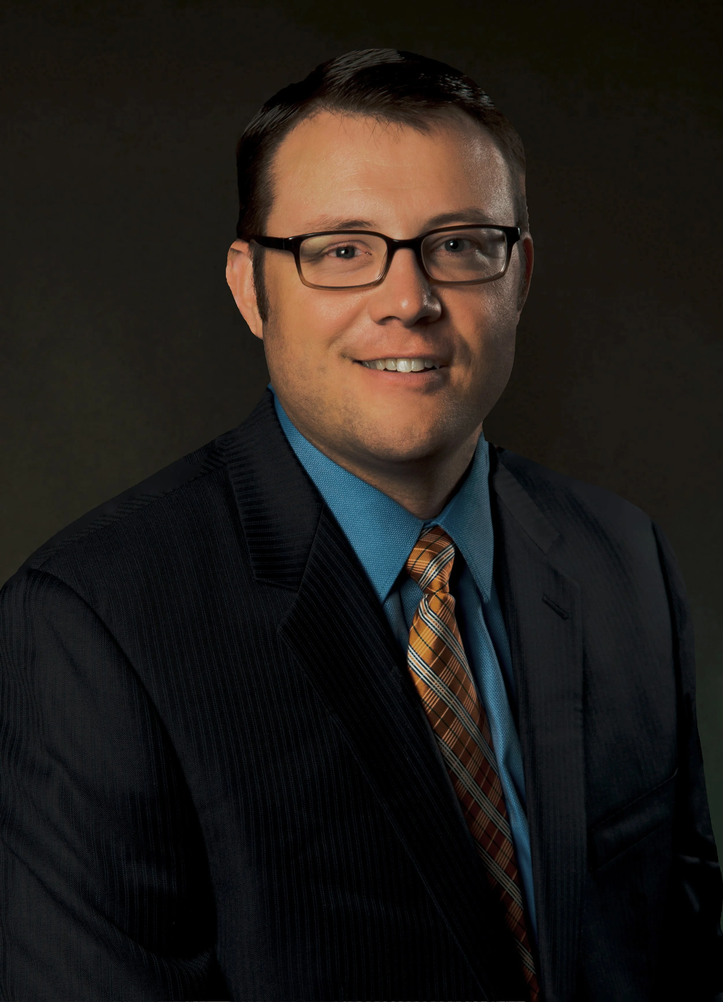 Professional portrait of a man in a suit with a checked orange tie, smiling against a dark background.
