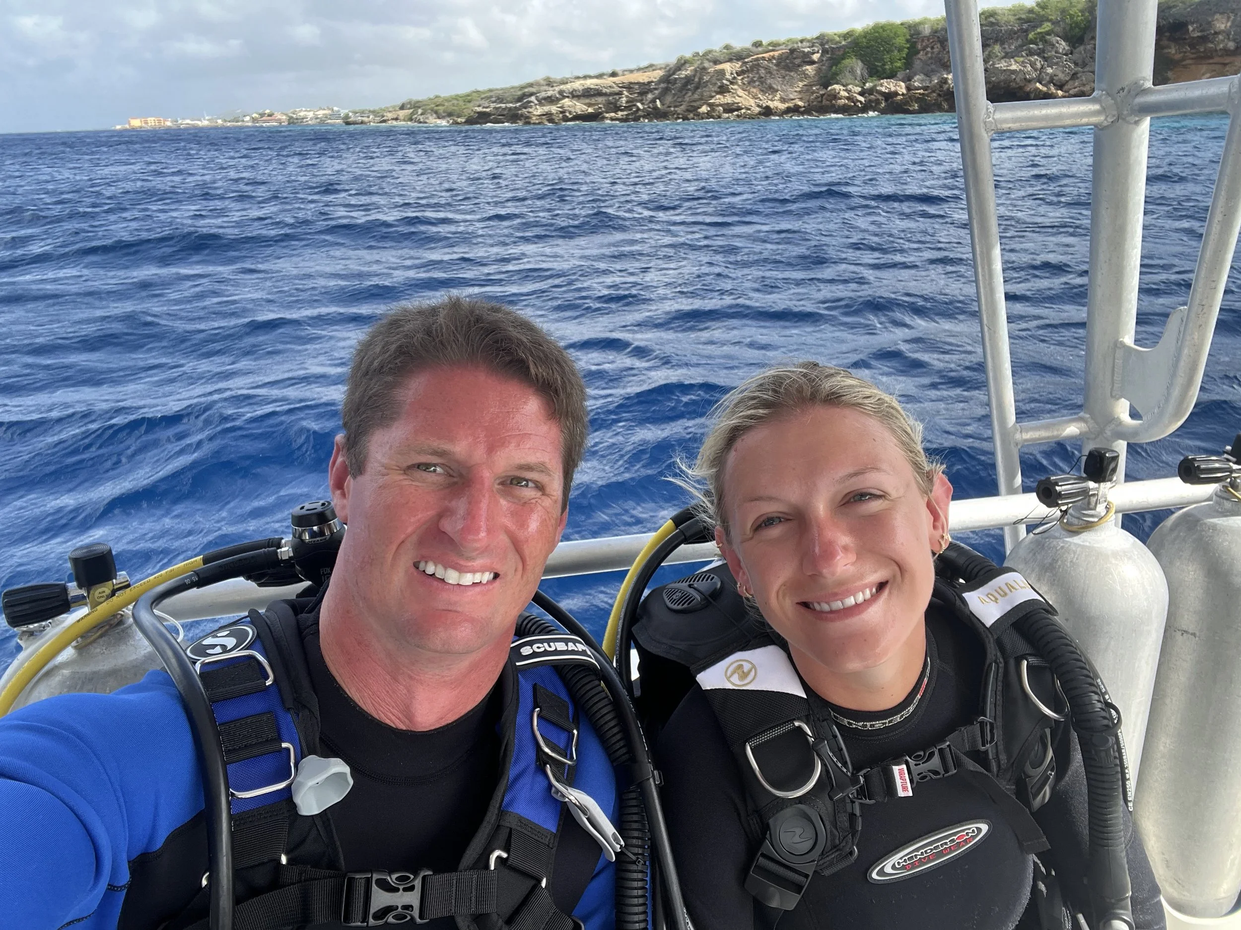 Two scuba divers taking a selfie on a boat with blue ocean water and a landmass with trees and buildings in the background.