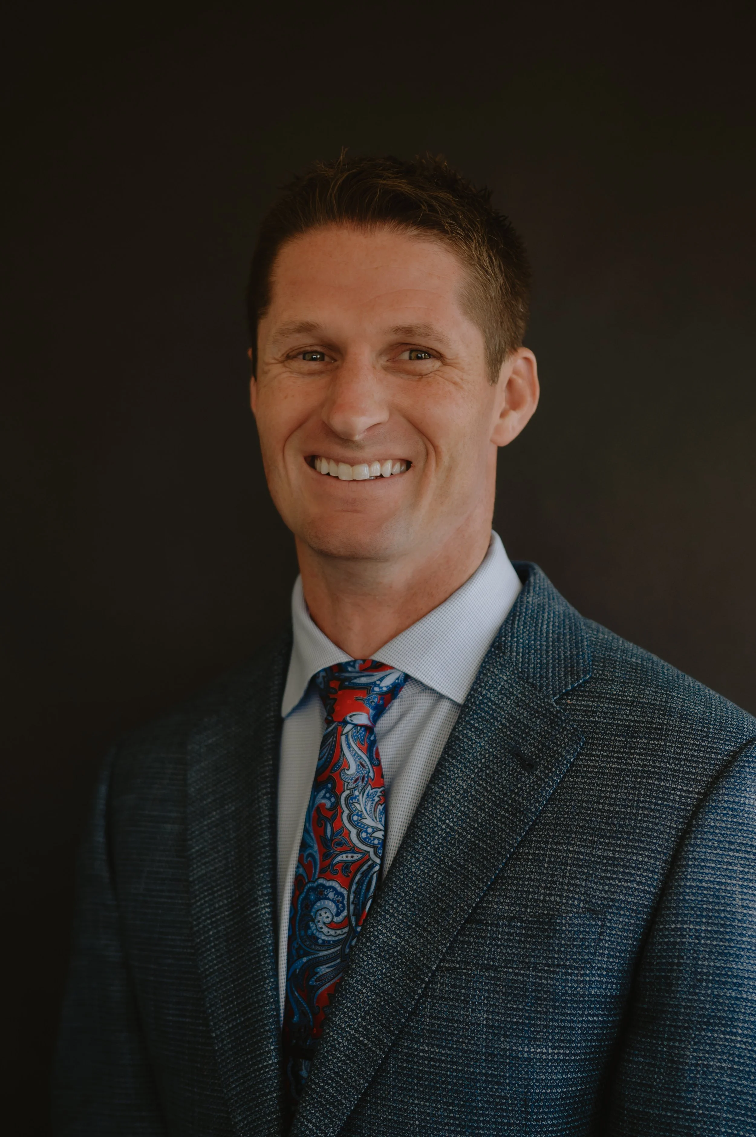 Headshot of a smiling man in a suit and tie against a black background.