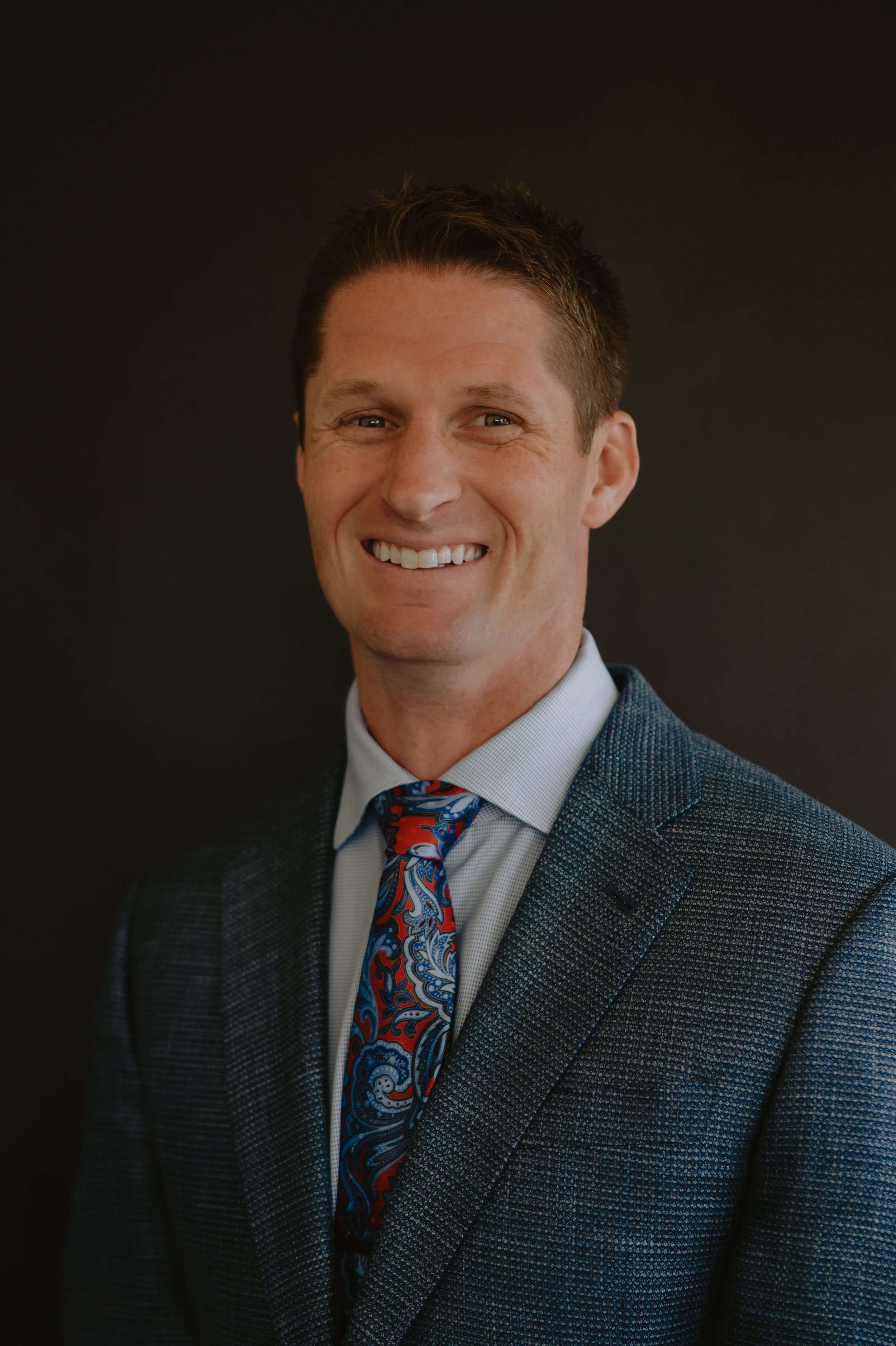 Headshot of a smiling man wearing a gray checkered blazer, light blue shirt, and a red, blue, and white paisley tie against a black background.