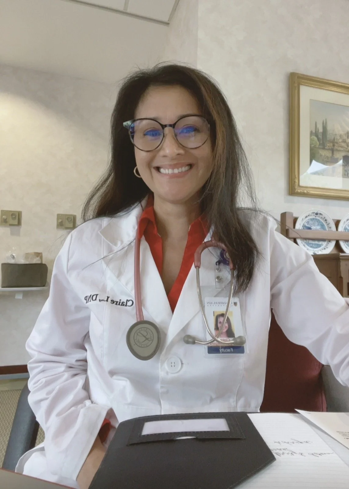 A woman wearing a white medical coat with a stethoscope around her neck sitting in an office or medical setting, smiling at the camera.