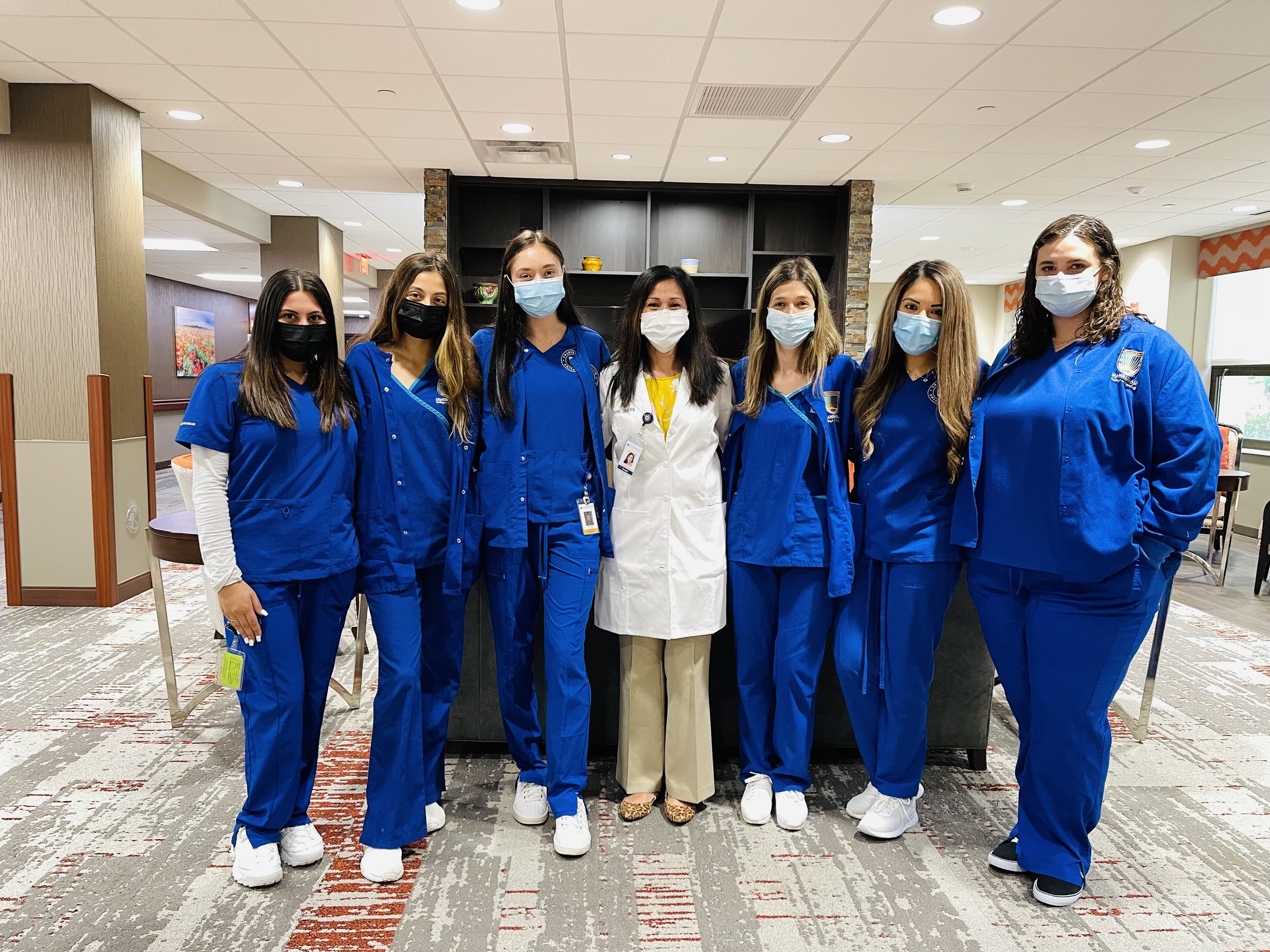 Group of seven women, six dressed in blue medical scrubs and one in white coat, standing in a hospital or healthcare facility, all wearing face masks.