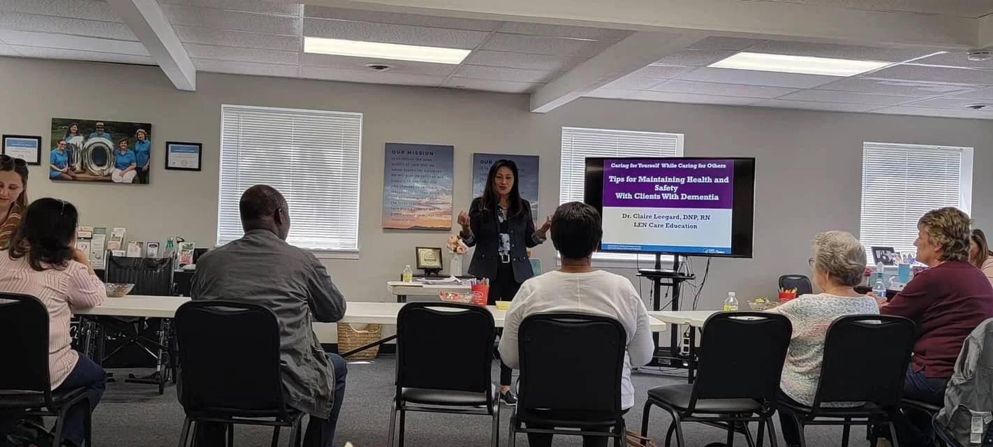 A woman standing and giving a presentation to a seated group of diverse adults. The presentation slide on the screen reads 'Tips for Maintaining Health and Safety With Clients With Dementia' and includes the presenter’s name, Dr. Claire Leegard, DNP, RN.