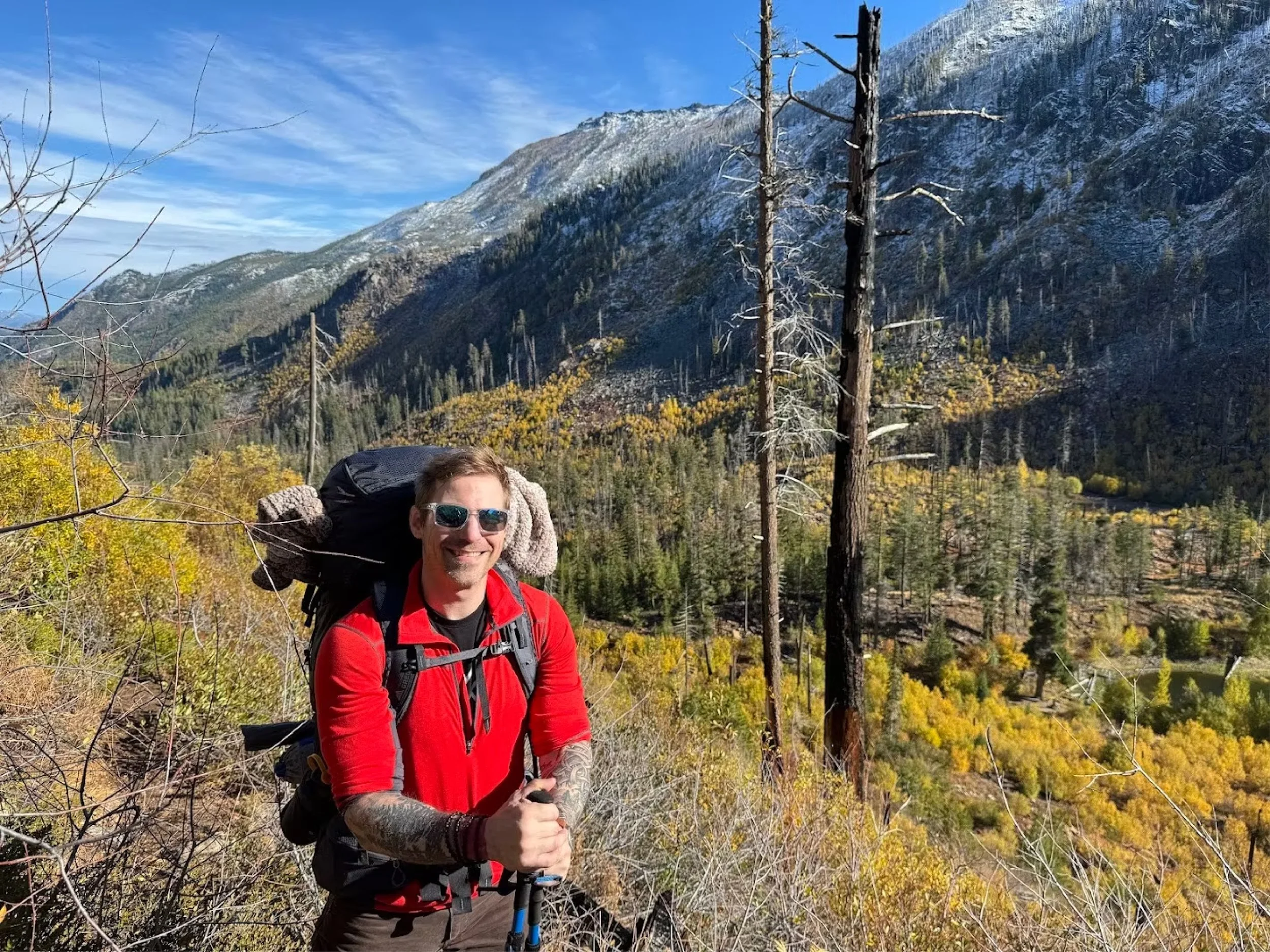A man in hiking gear standing in a mountain landscape with pine trees, a valley, and snow-capped mountains in the background on a sunny day.