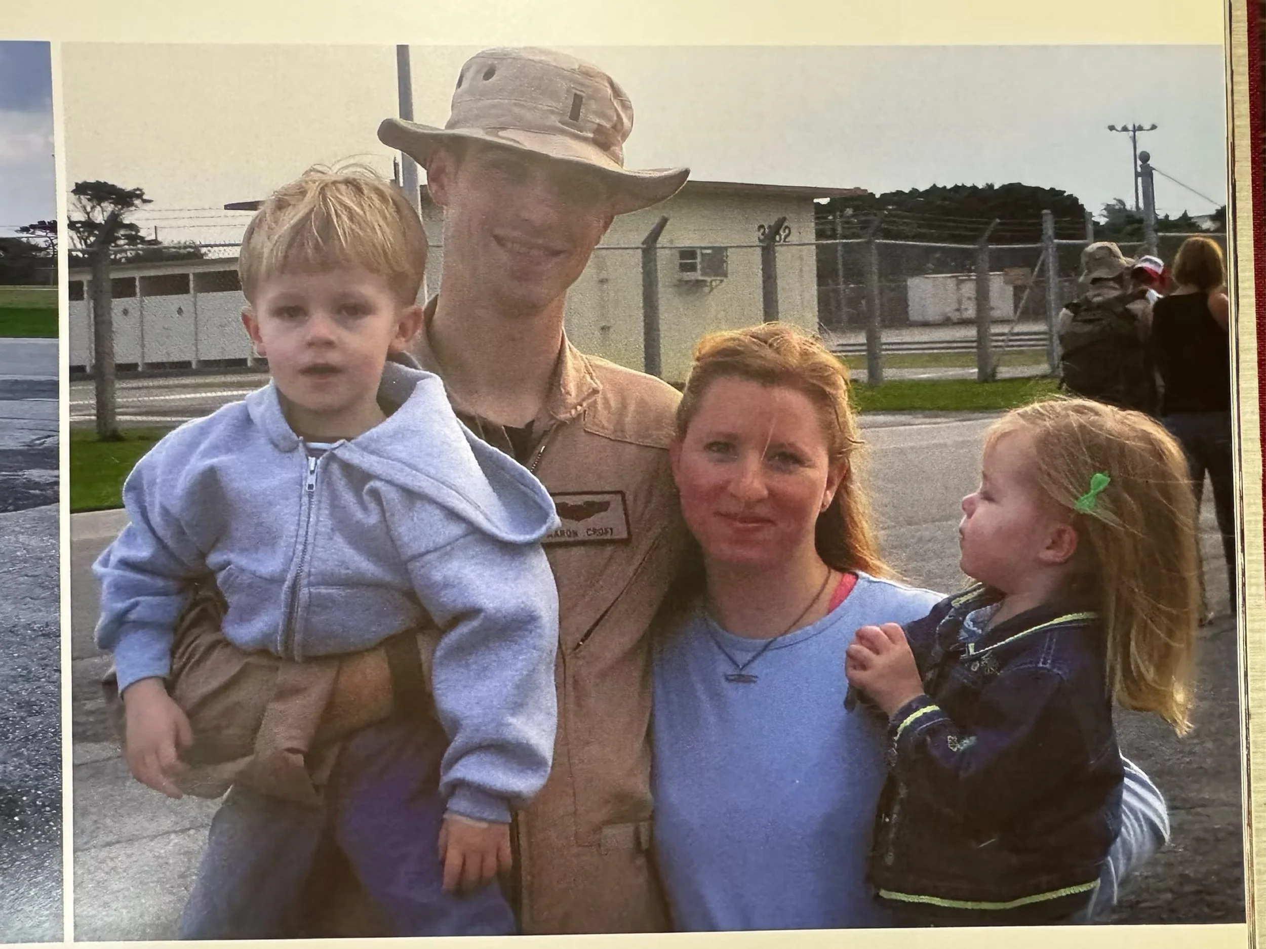 A family of four outdoors, with a man in military uniform holding a young boy, a woman with red hair, and a young girl with a green hair clip.