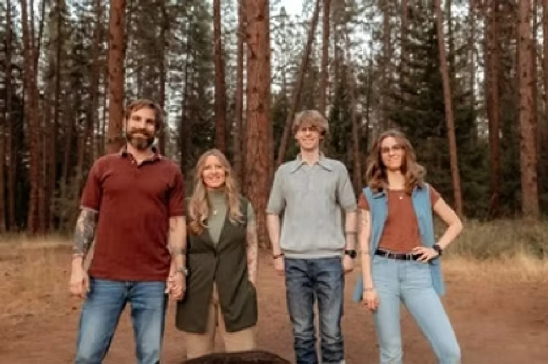 A group of four young adults standing in a forest, smiling at the camera, with tall trees in the background.