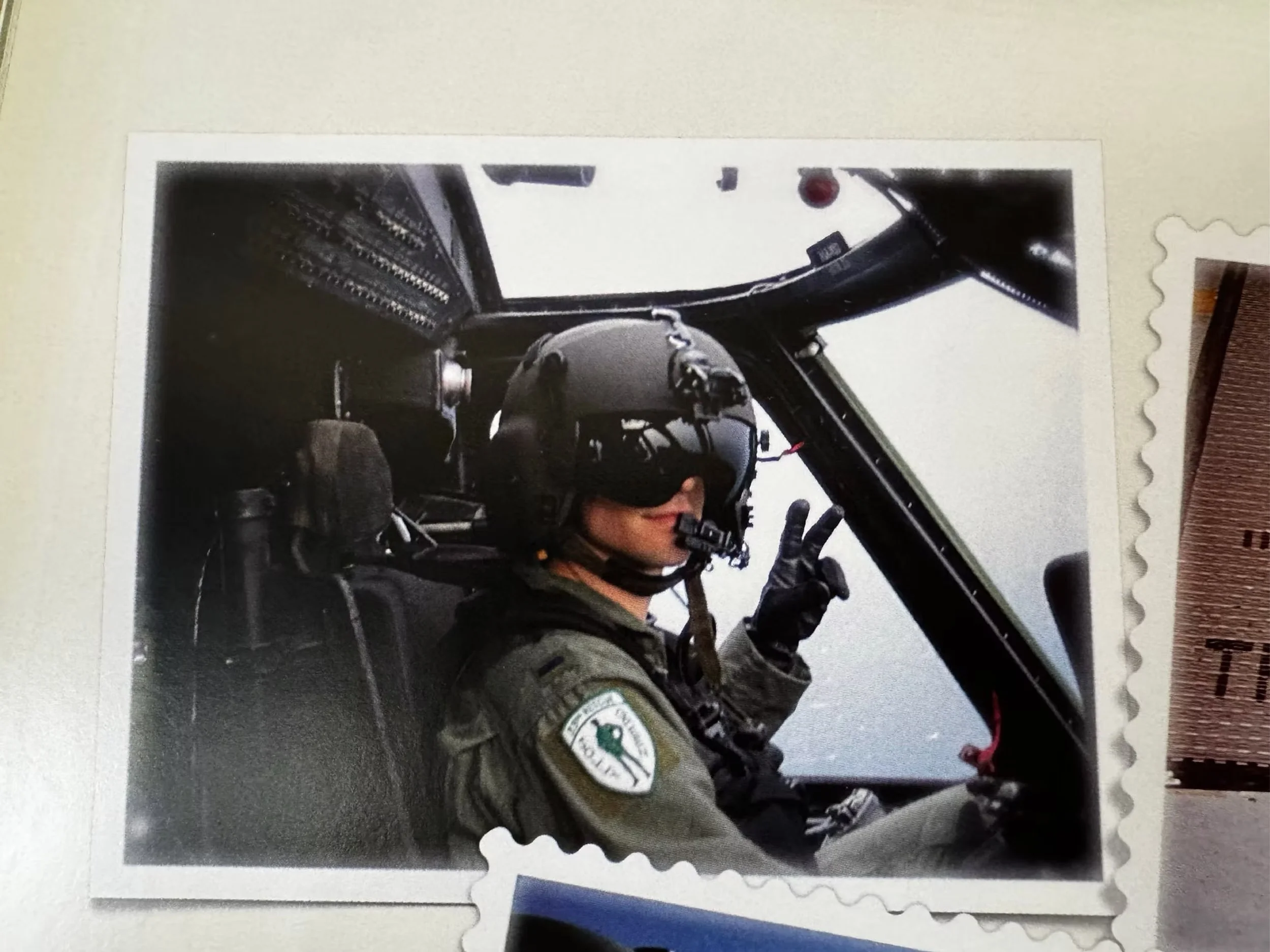 A pilot sitting inside a fighter jet cockpit, wearing a helmet and visor, making a peace sign with their hand.