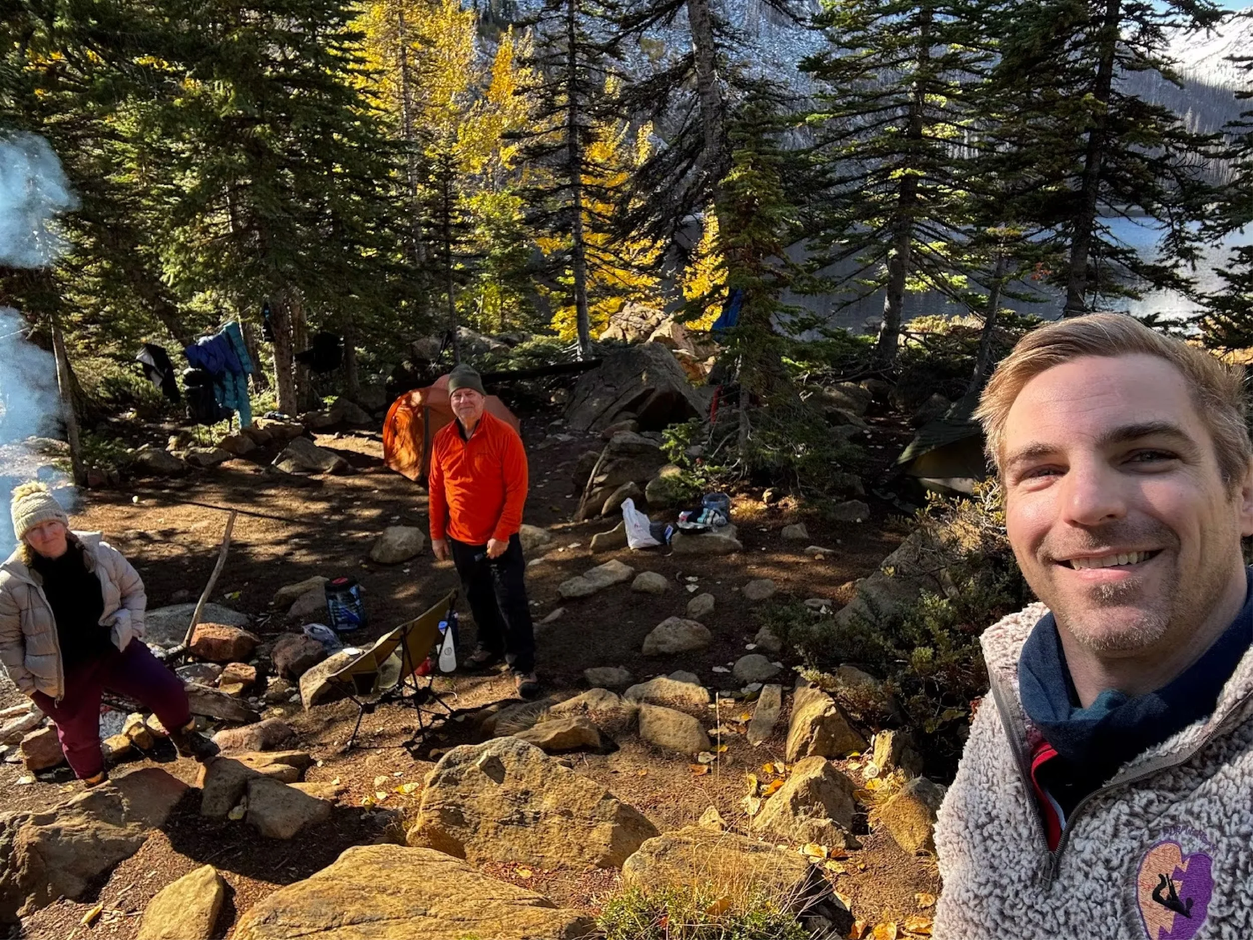 Three people camping in a wooded area with tall evergreen trees, rocks, and a tent in the background. One person is taking a selfie, two others are standing near camping gear and a campfire.