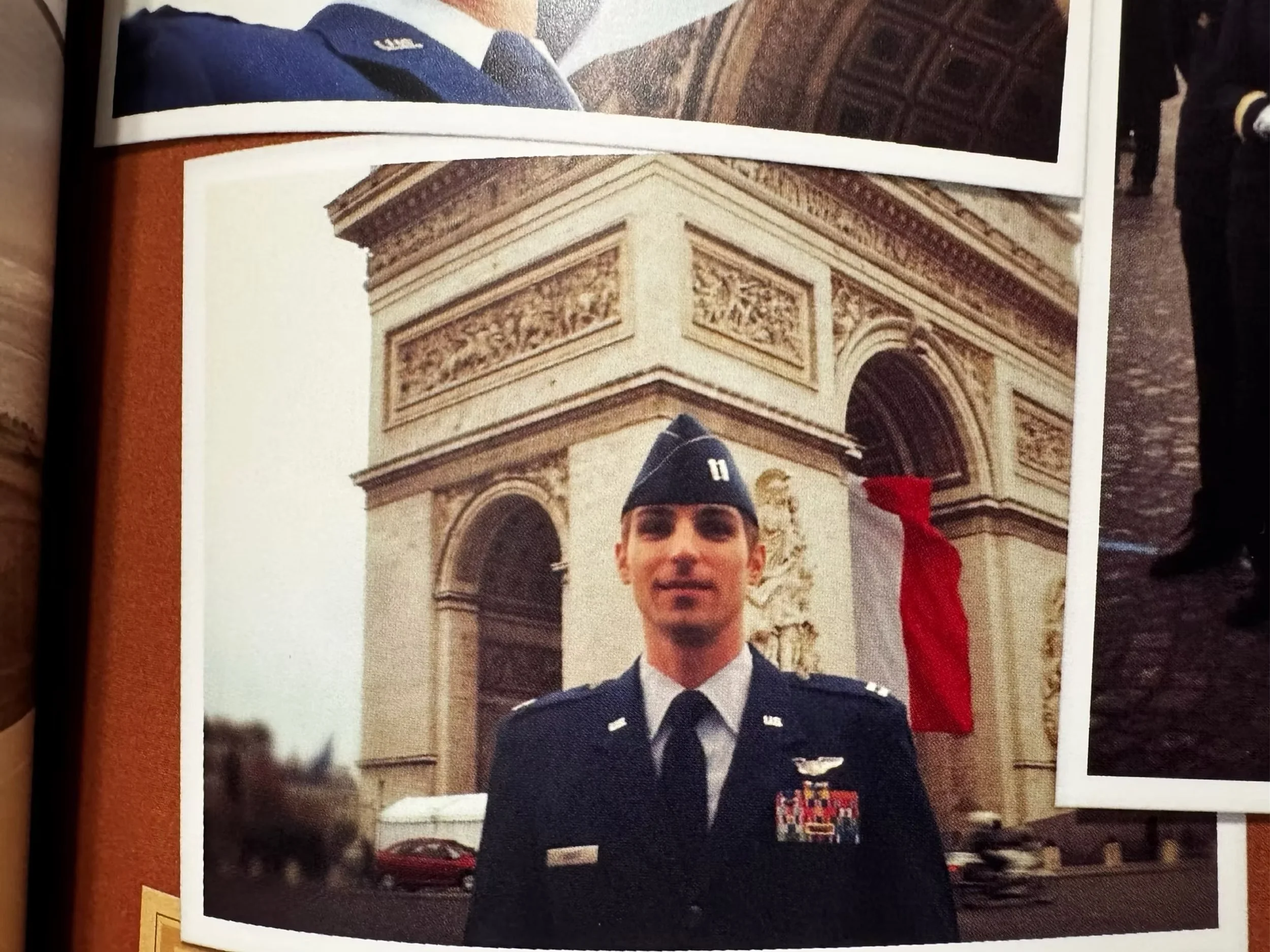 A man in military uniform standing in front of the Arc de Triomphe in Paris, France, with a red cloth or decoration hanging on the monument.