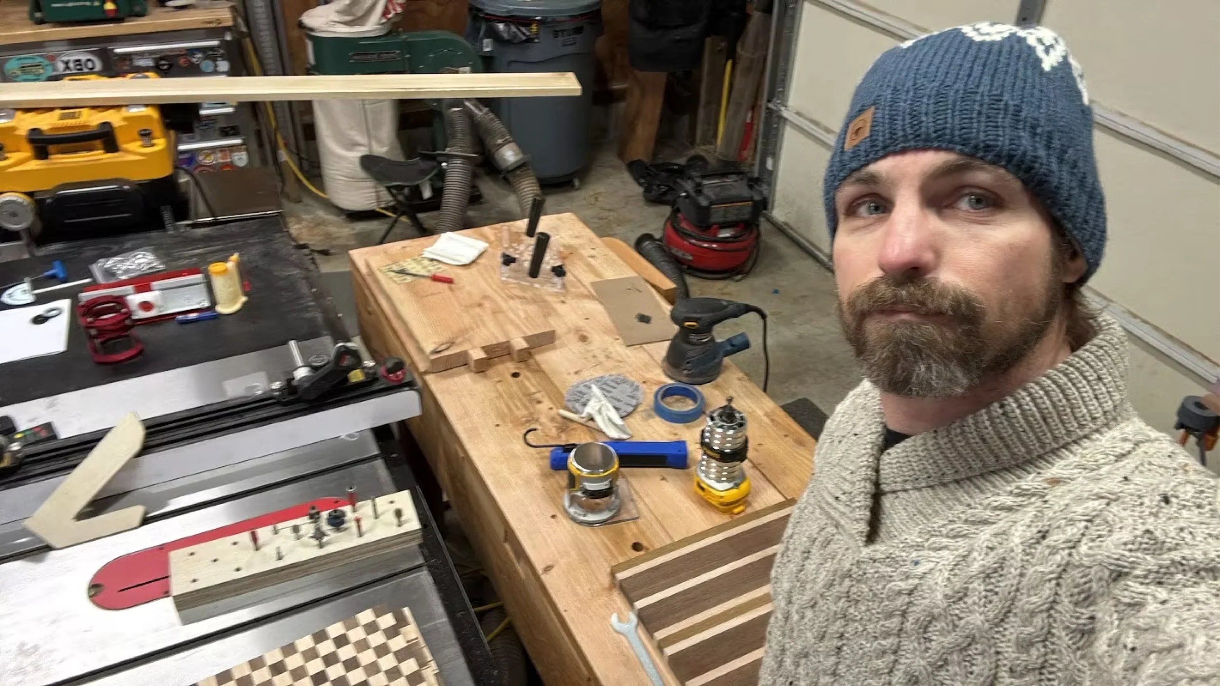 A man with a beard wearing a blue knit cap and a gray sweater standing in a woodworking workshop. The workshop contains a workbench with tools and materials, such as clamps, a flashlight, a sander, and pieces of wood, including a chessboard pattern.