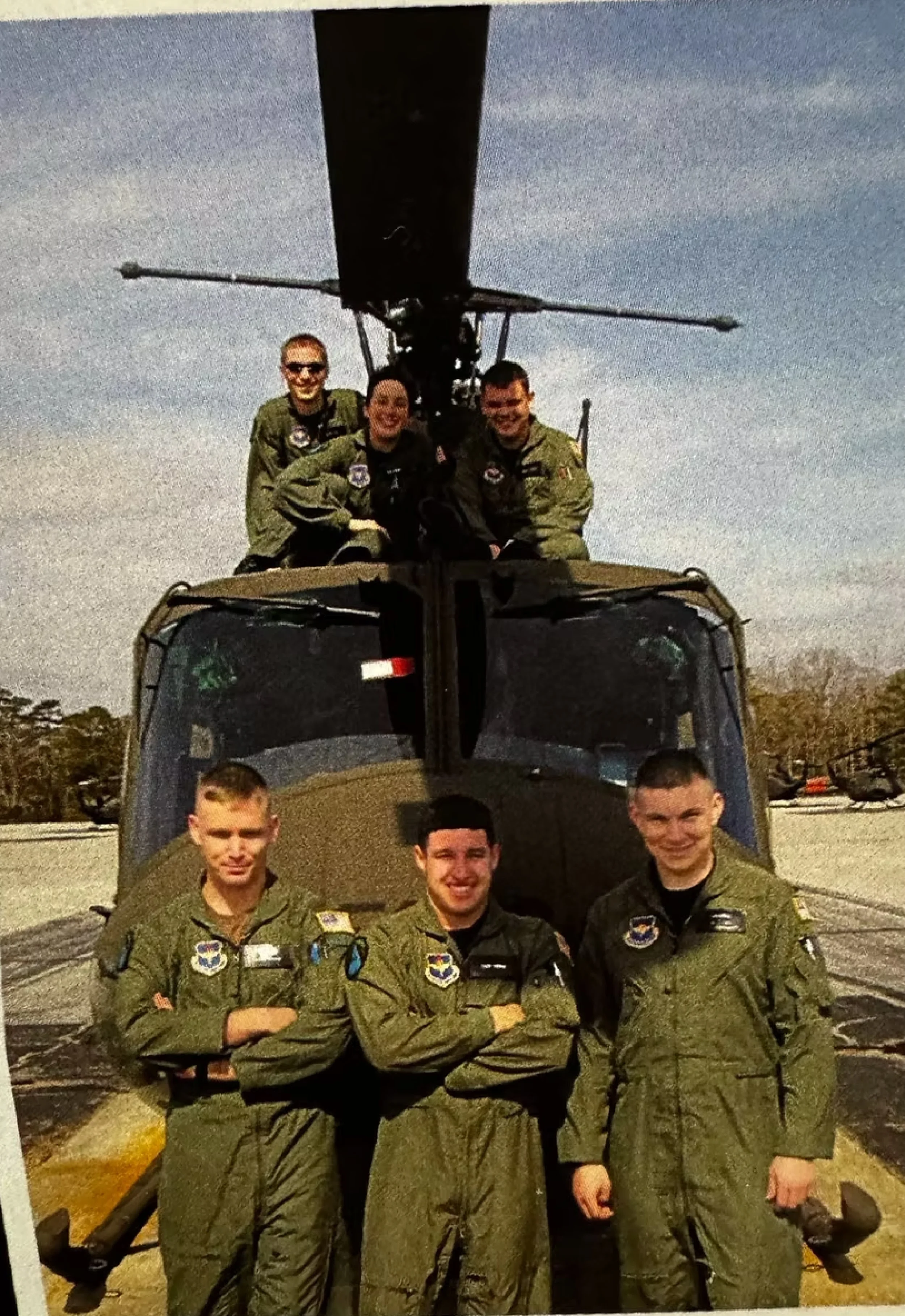 Six people in military flight suits, three standing in front, and three sitting on top of a helicopter, outdoors on a sunny day.