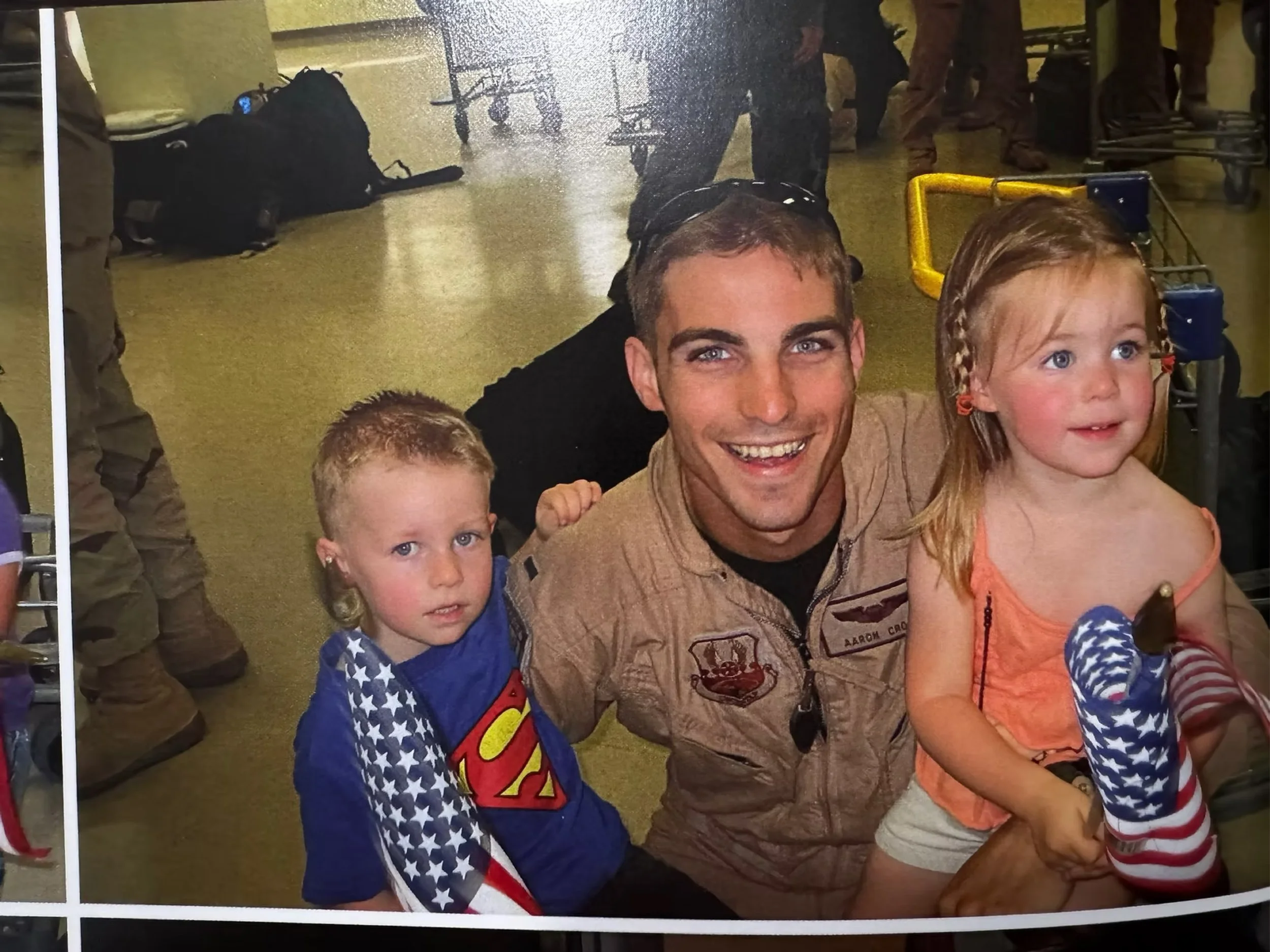A man in military uniform with two young children, a boy and a girl, sitting on the floor at an airport with luggage and other travelers in the background.