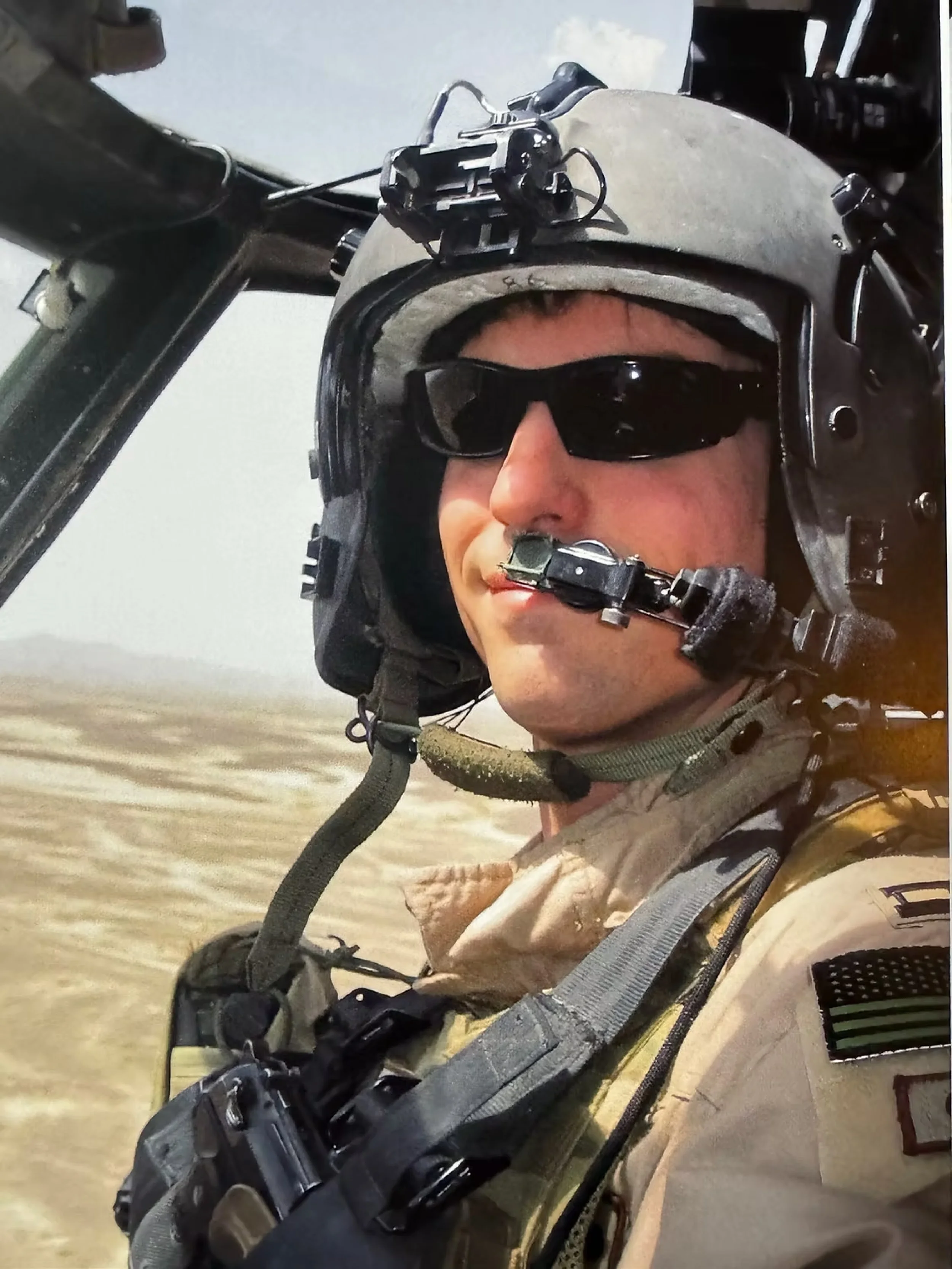 Close-up of a military pilot in flight gear, wearing a helmet with mounted equipment and sunglasses, sitting in the cockpit with a desert landscape visible outside.