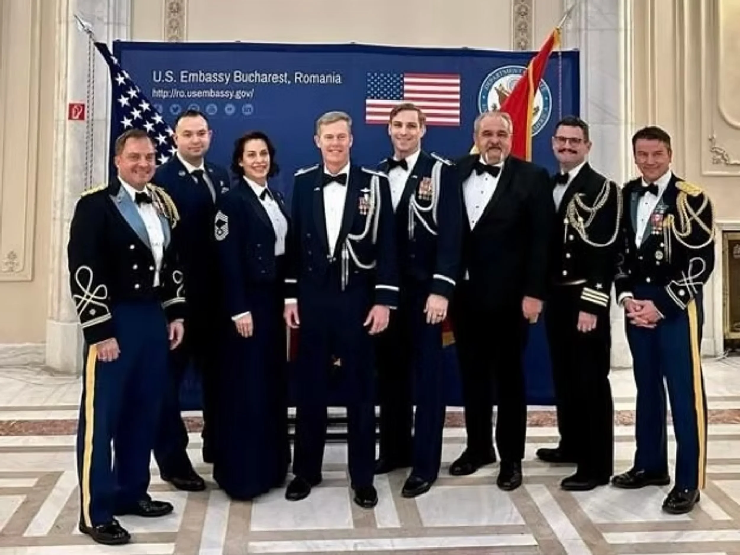 Group of nine people in military formal uniforms posing in front of a U.S. Embassy banner and flags in a decorated indoor setting.