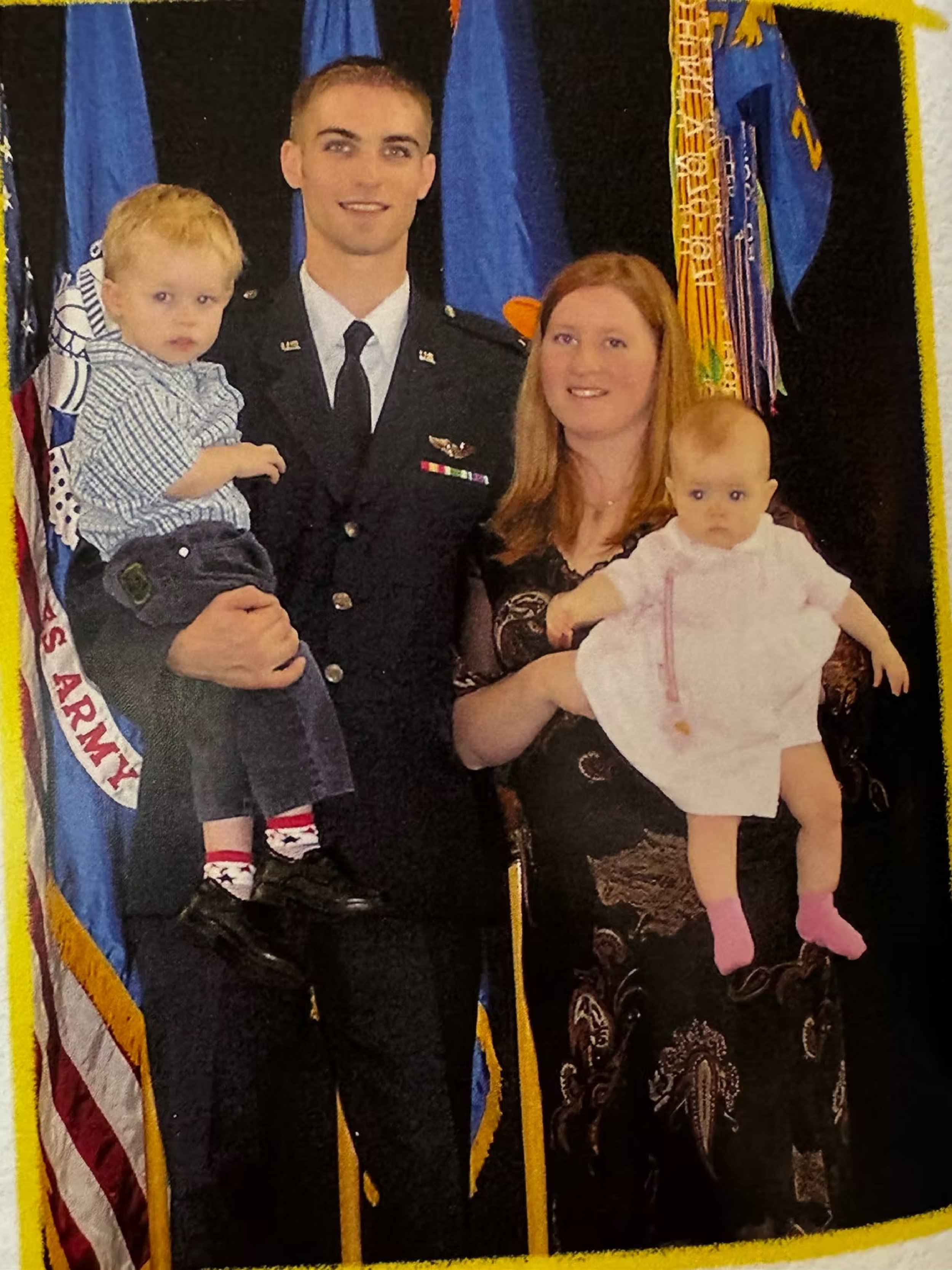 A family photo with two adults and two young children, with flags in the background, including one that reads 'U.S. Army,' suggesting a military or patriotic setting.