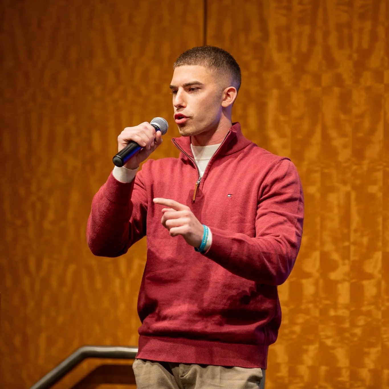 A young man with short hair, wearing a red pullover with a quarter zip and a beige shirt underneath, speaking into a microphone on stage with a wooden backdrop.