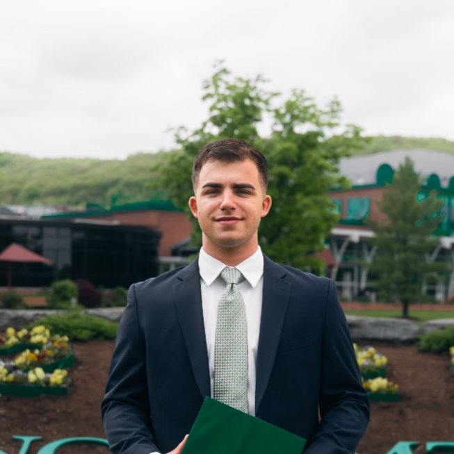 Young man in a dark suit, white shirt, and light tie holding a green folder outdoors in front of a garden and modern buildings.