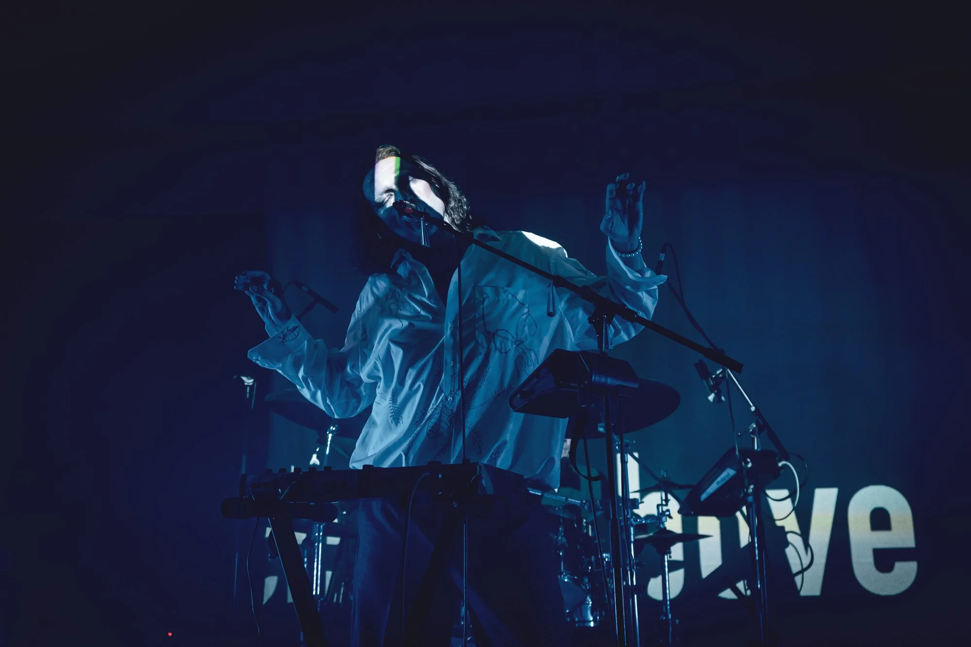 A musician performing on stage at night, singing into a microphone and playing electronic music equipment, with a dark background and blue lighting.
