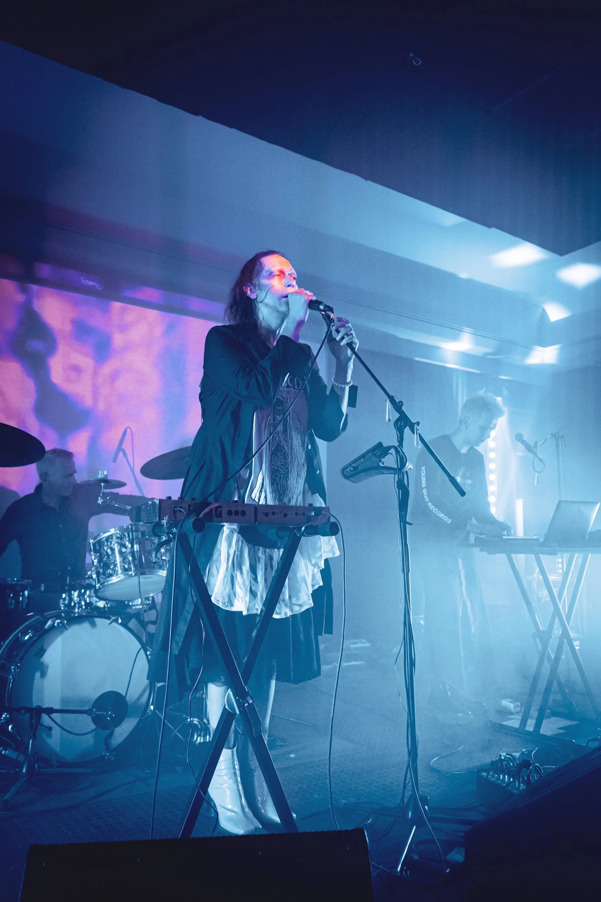 A band performs on stage, with a female singer at the front holding a microphone, a drummer in the back, and a keyboardist to the right, under colorful stage lighting.