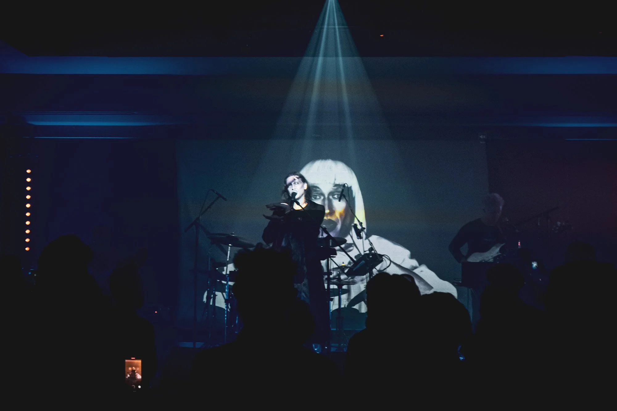 A live music performance on stage with a woman singing into a microphone and a guitarist playing beside her, with a large black and white image of a woman's face projected on the backdrop.