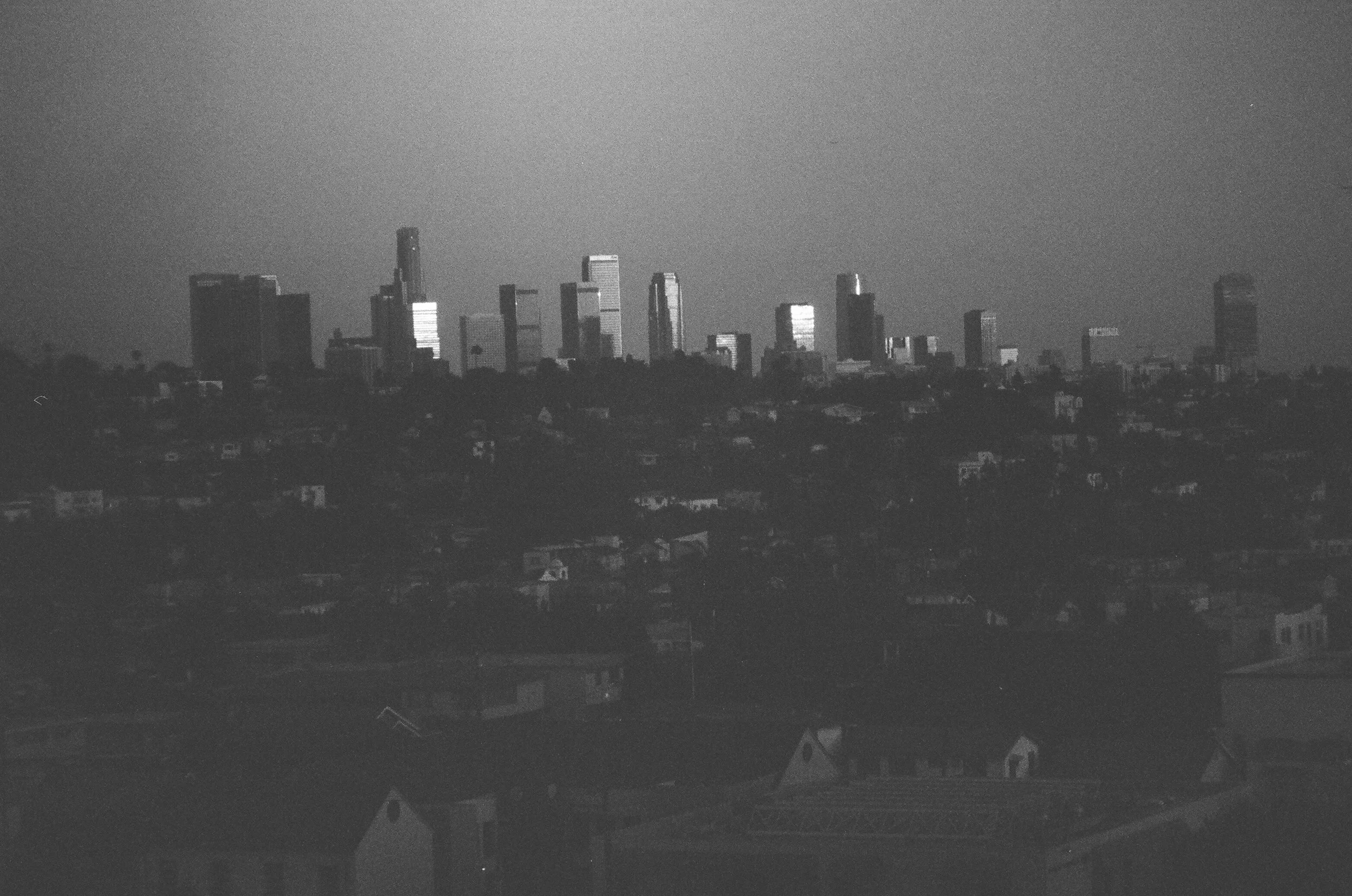 Black and white photo of a city skyline with tall buildings and skyscrapers, seen from a distance across a residential area, under a clear sky.