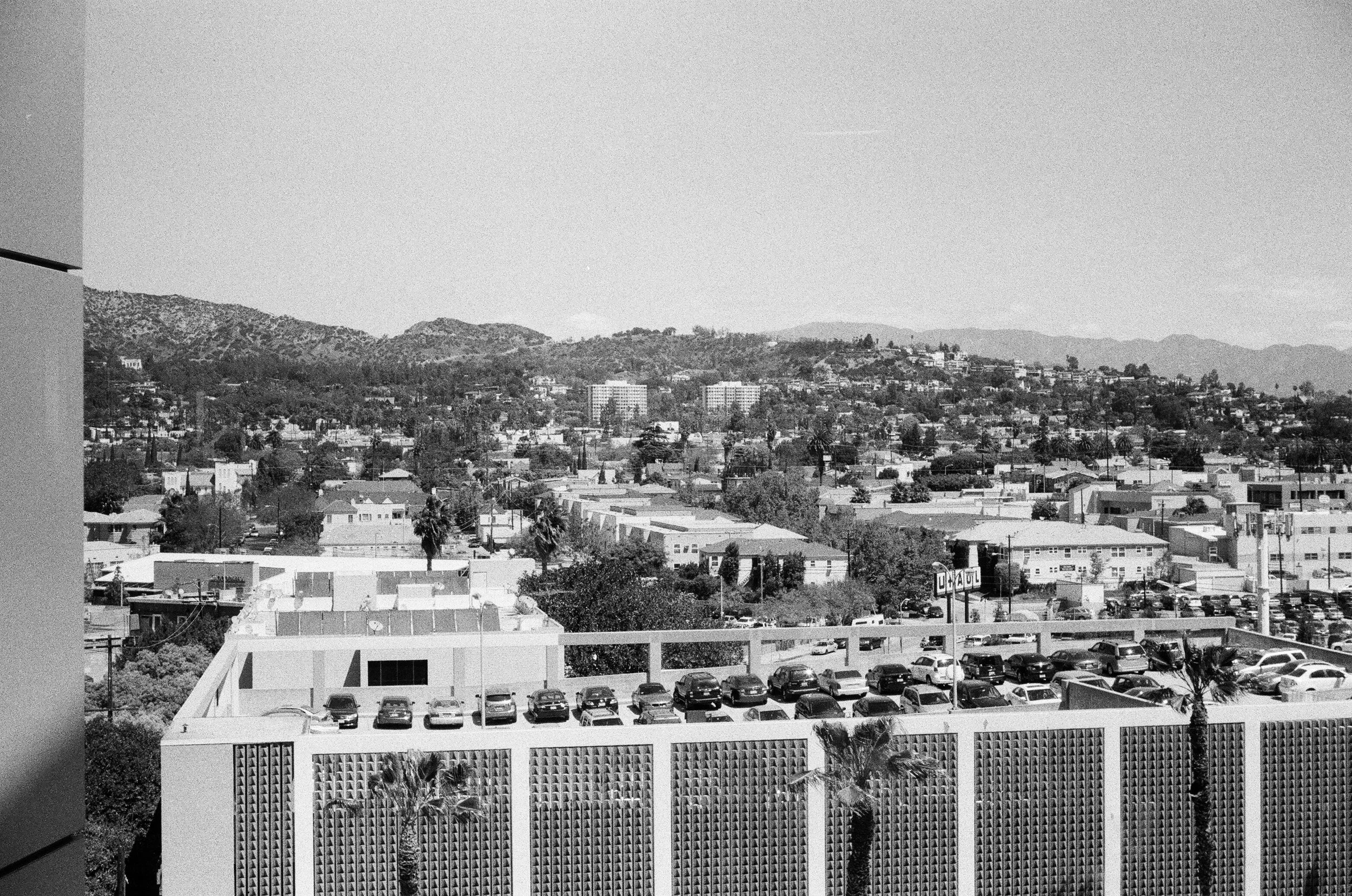 Black and white photograph of a downtown cityscape with hills or mountains in the background. Foreground features a modern building with a perforated facade, a parking lot with several cars, and a white railing. Midground has various buildings, and t