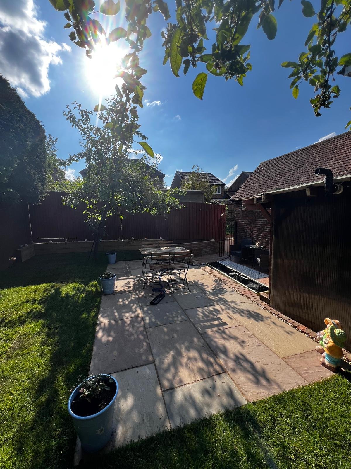 A backyard patio with a sunlit tree, potted plants, a table and chairs, and a small gnome statue, under a bright blue sky with some clouds.
