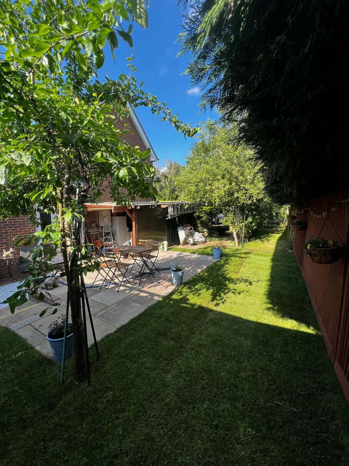 A backyard with a grassy lawn, potted plants, and a stone patio with outdoor furniture. There are trees and bushes, and a blue sky with some clouds overhead.