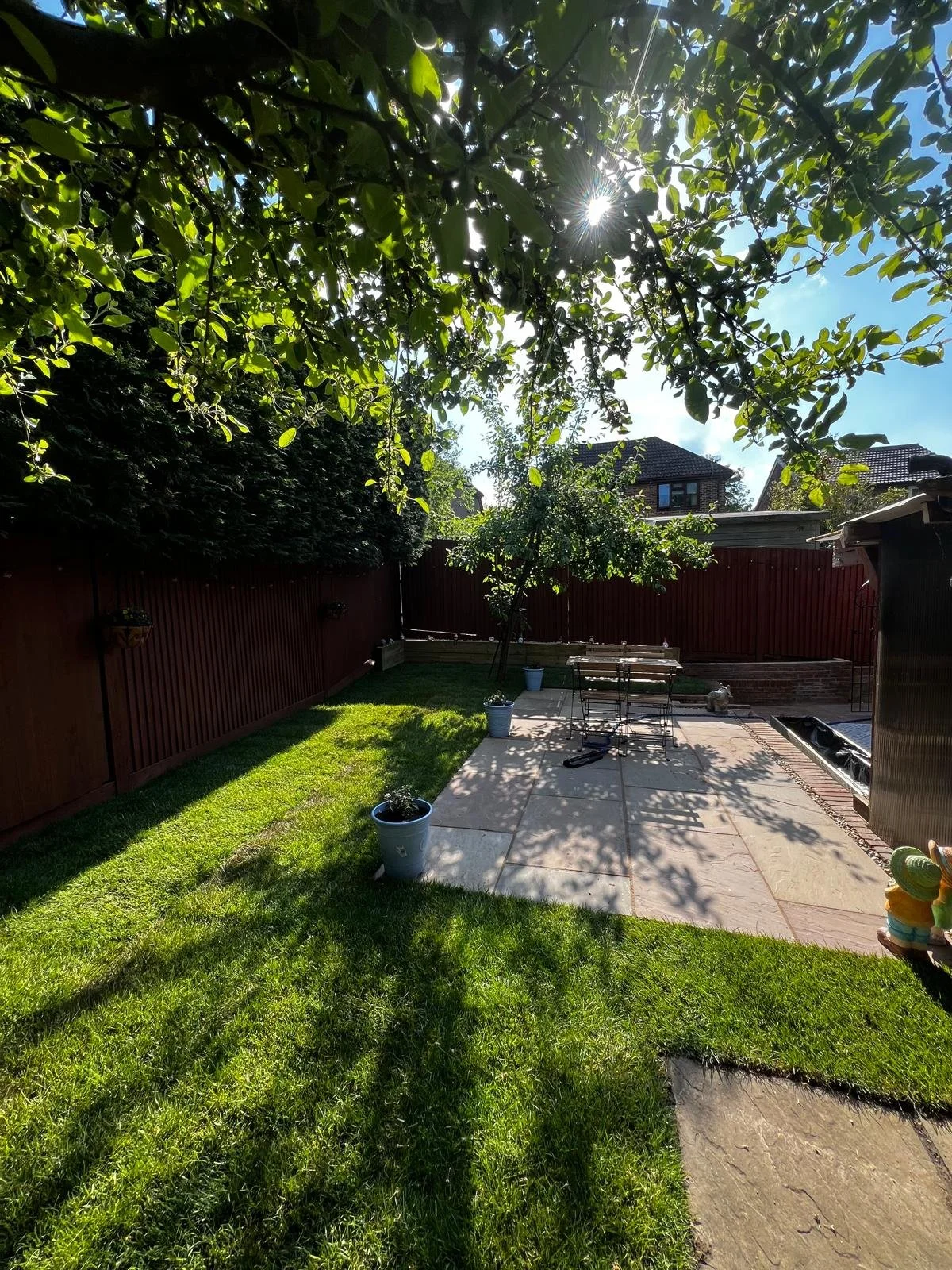 A backyard with a grassy area and a patio, shaded by large trees with sunlight filtering through the leaves, and a red wooden fence in the background.