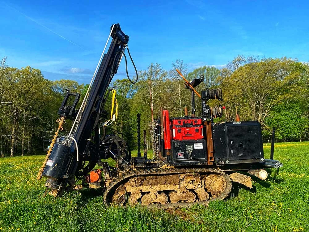 A tracked post knocking machine with a tall black arm and hydraulic drill attached, on green grass with trees and a blue sky in the background.