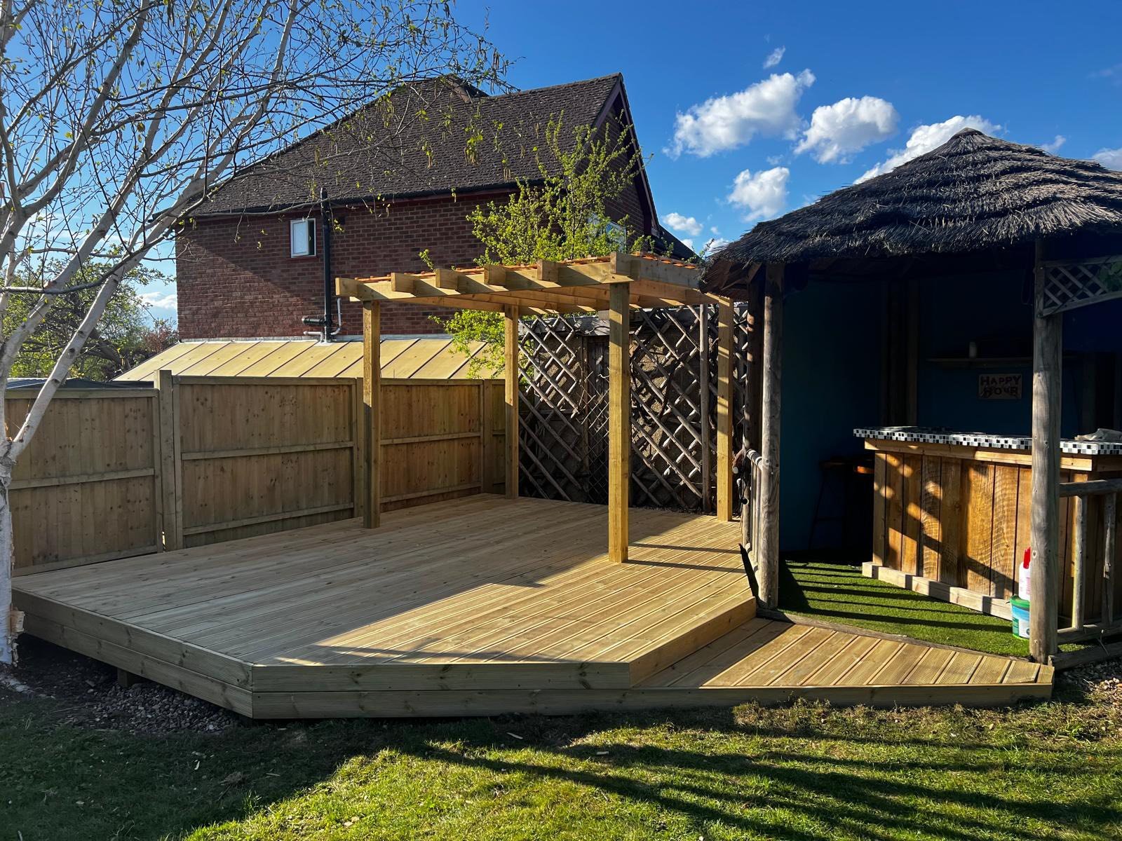A backyard patio with a wooden deck, partially under construction, featuring a pergola frame, a wooden fence, a small wooden structure with a thatched roof, and a grassy area with trees and a house in the background.