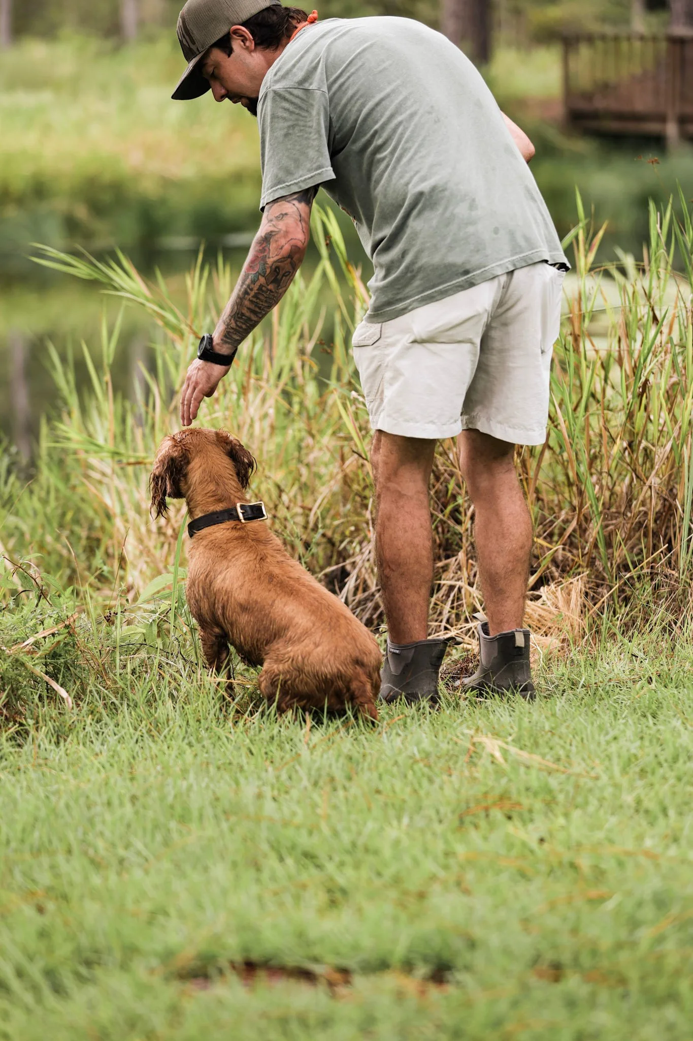 A man with tattoos, wearing a gray t-shirt, beige shorts, and black boots, is kneeling and petting a small brown dog by a pond, in a grassy and wooded area.
