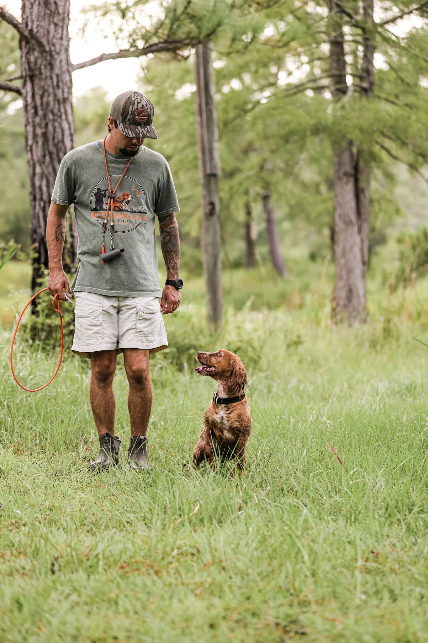 A man standing outdoors with a dog sitting attentively on the grass, trees in the background.