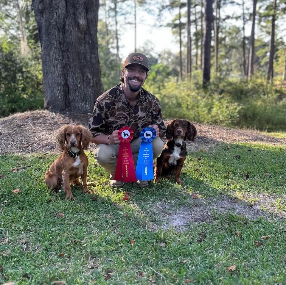A smiling man in camouflage shirt and cap kneels on grass, holding two award ribbons, with two dogs sitting on either side. The dogs are posing outdoors near a tree in a park-like setting with trees and greenery in the background.