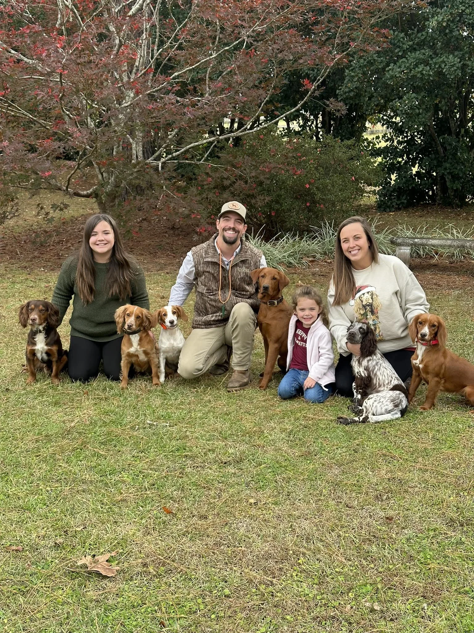 A family of five people, including two adult women, one adult man, and two young girls, posing outdoors on grass with nine dogs of various breeds and sizes, under trees with fall foliage.