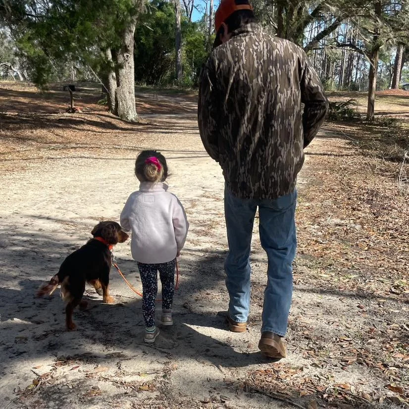 A man, a young girl, and a dog walking on a dirt path in a wooded area during daytime. The girl is holding a leash attached to the dog.