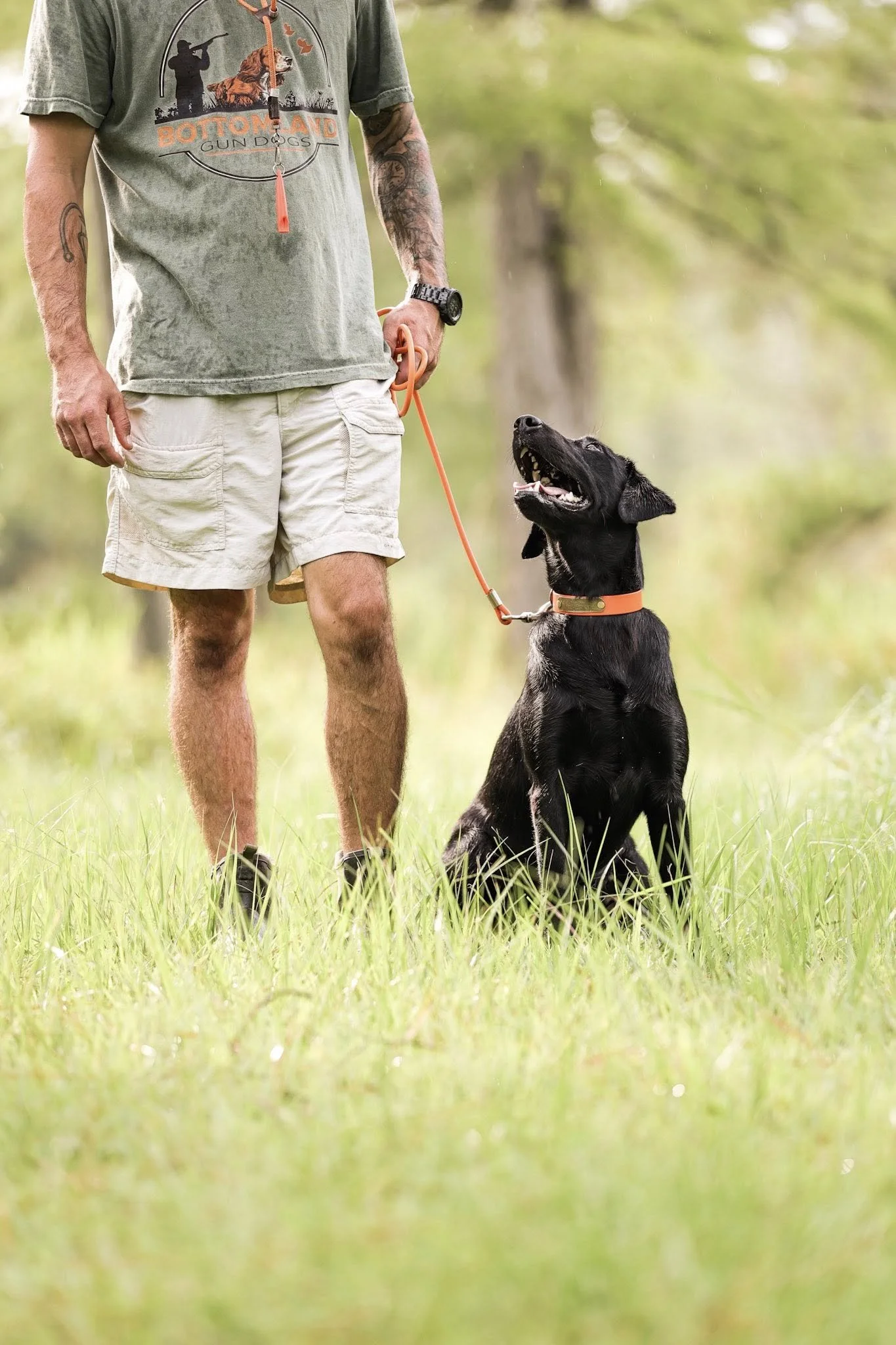 A man standing on a grassy field with a black dog sitting obediently by his side. Both are outdoors with trees in the background. The man is wearing a gray t-shirt with a graphic, beige shorts, and has tattoos and a watch on his left wrist. The dog i