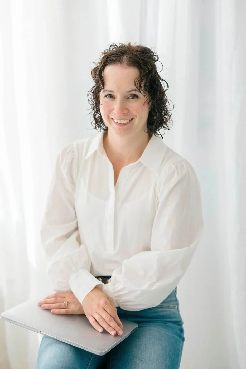 Jileah, Online Business Manager: a woman with curly dark hair smiling, wearing a white blouse and blue jeans, holding a closed laptop, sitting in front of white curtains.